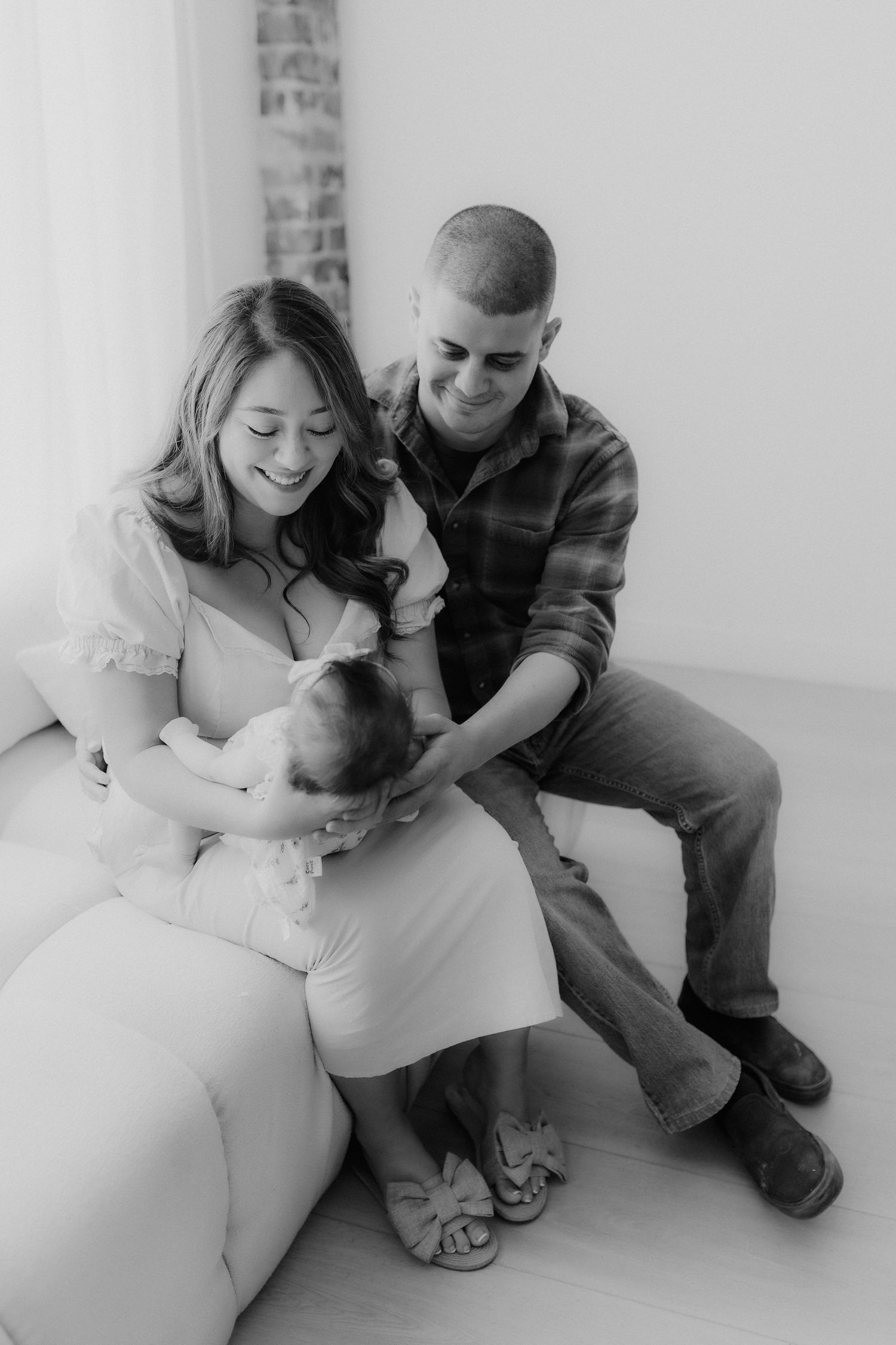 Black and white photo of smiling young family with newborn baby sitting together on white couch.