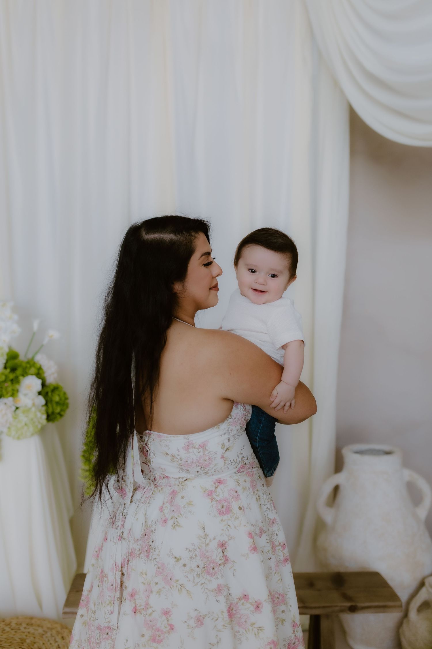 Mother in floral dress holding smiling baby boy in white studio setting with draped curtains and flowers.