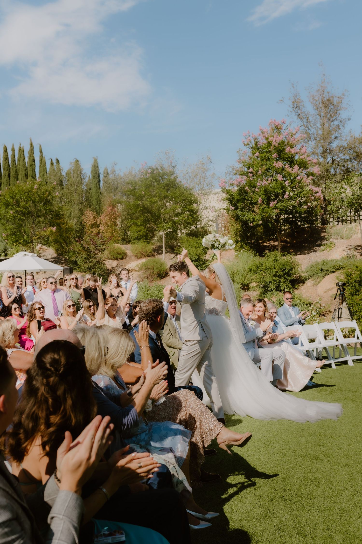 Newlyweds celebrate joyfully as guests cheer and applaud during outdoor wedding ceremony exit.