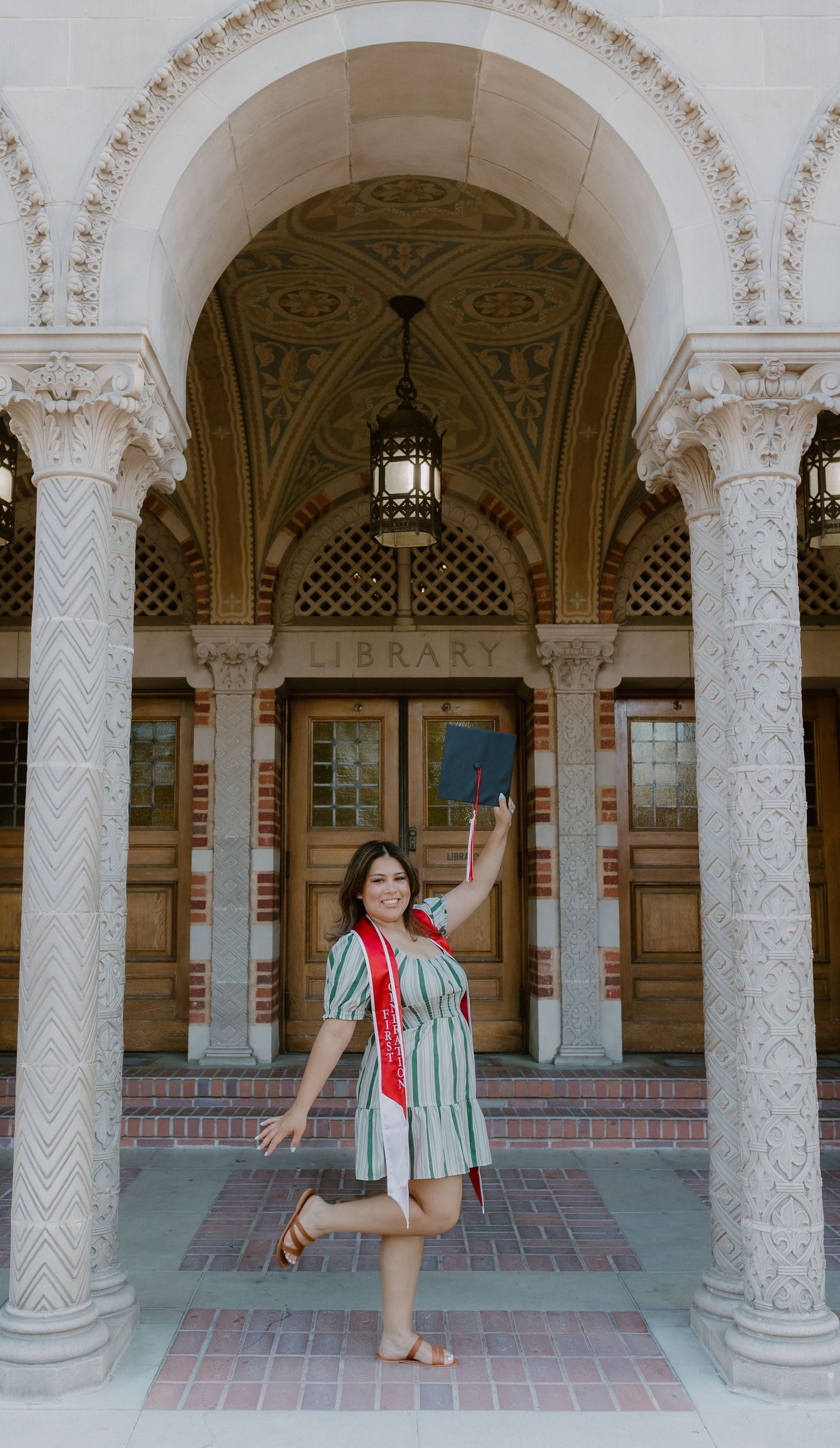 Smiling graduate in striped dress raises cap outside ornate university library with decorative archway. Fresno, California