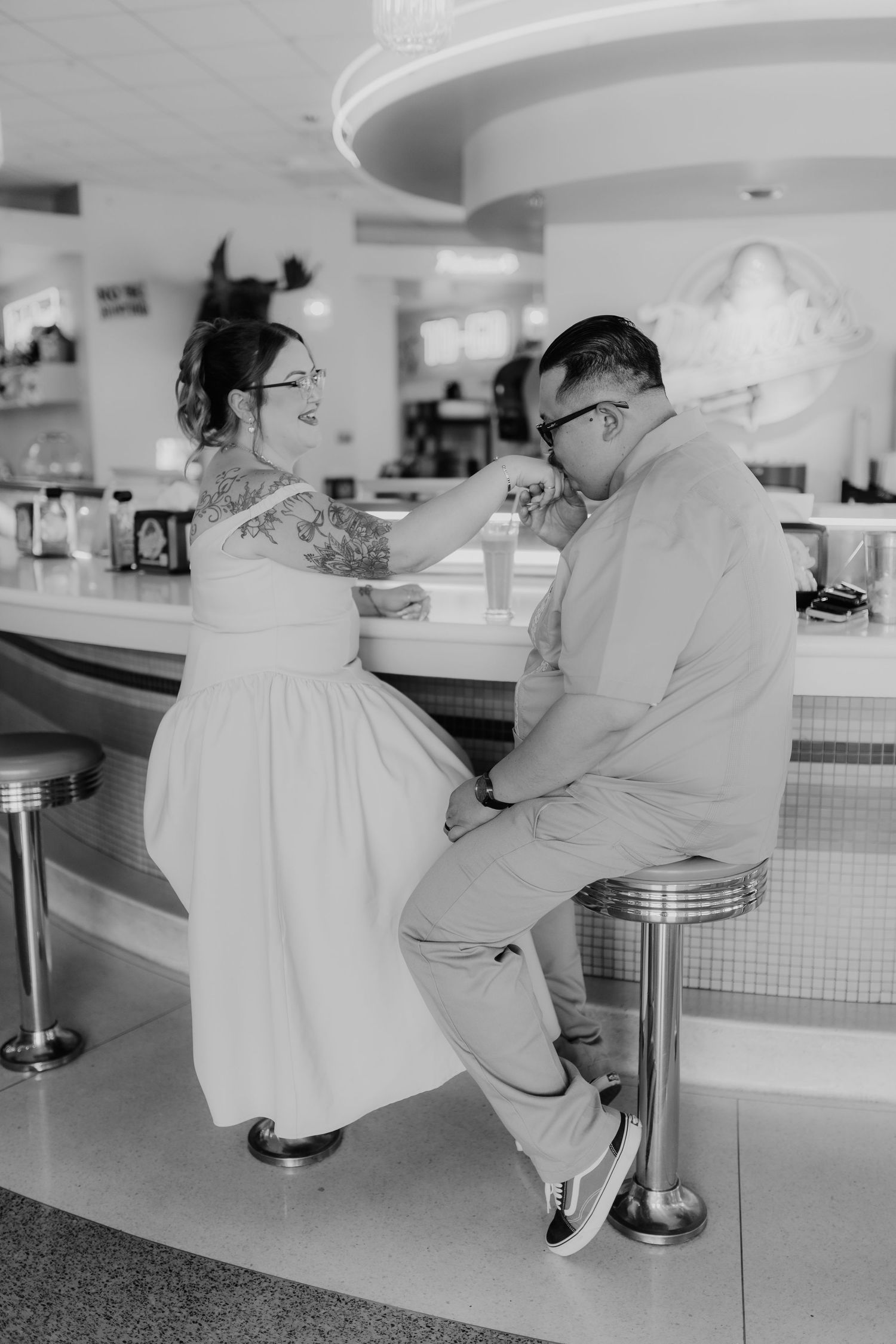 Black and white photo of couple sharing milkshake at retro diner counter, bride in white dress with tattoos.