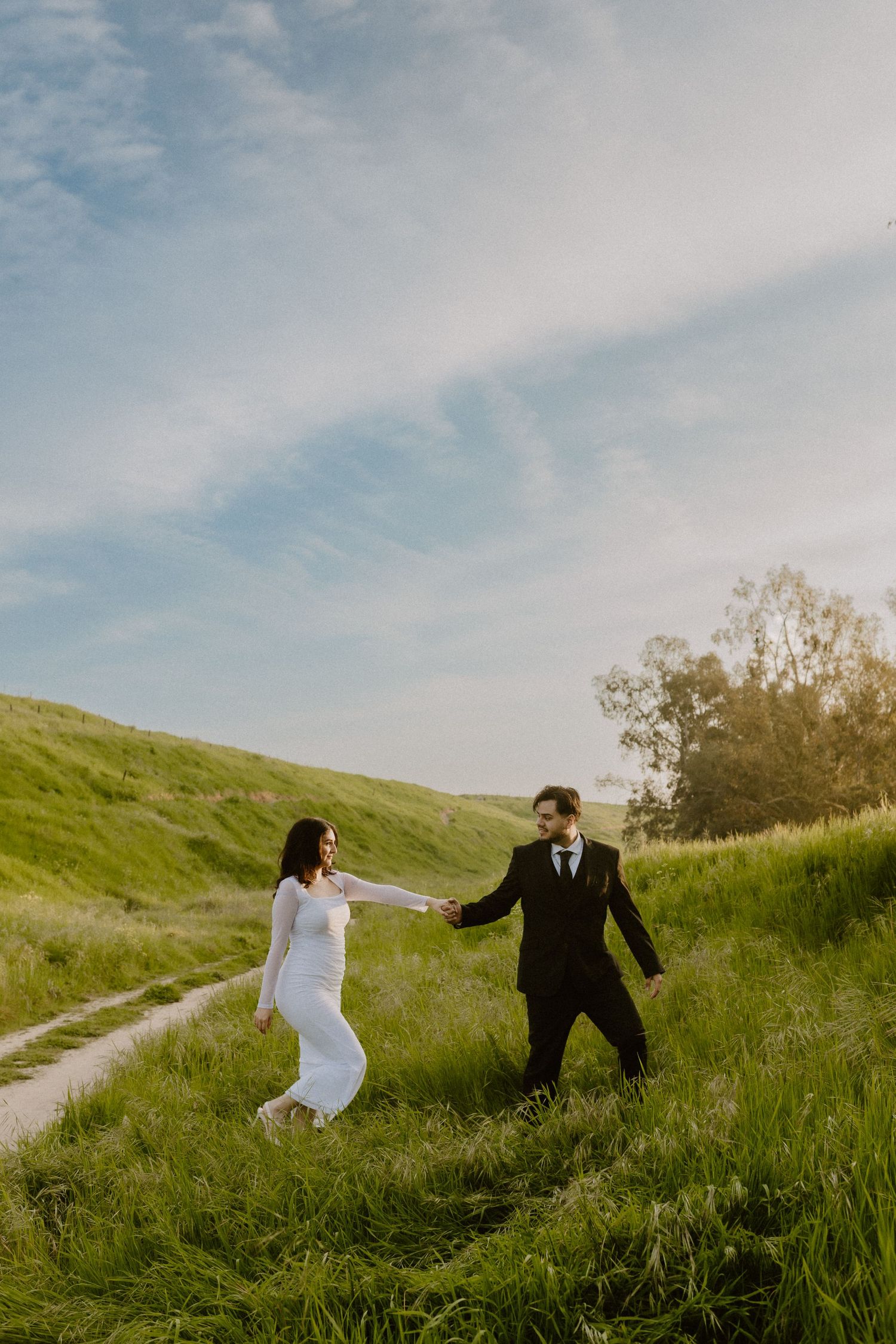 Bride and groom in wedding attire holding hands while walking on green hillside path.