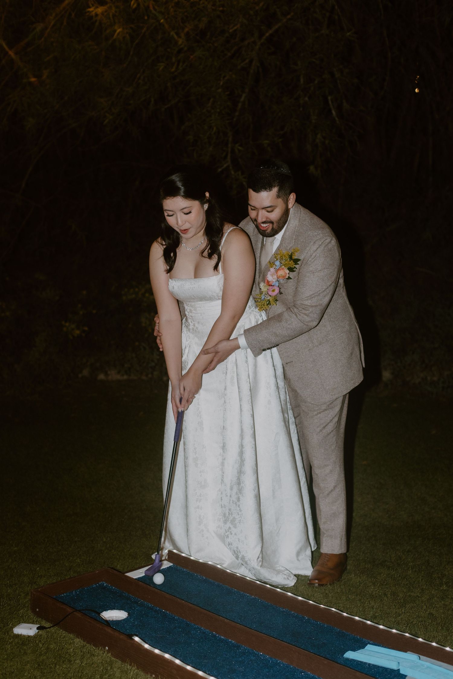 Groom helping bride putt during mini golf wedding reception activity at night outdoor event.