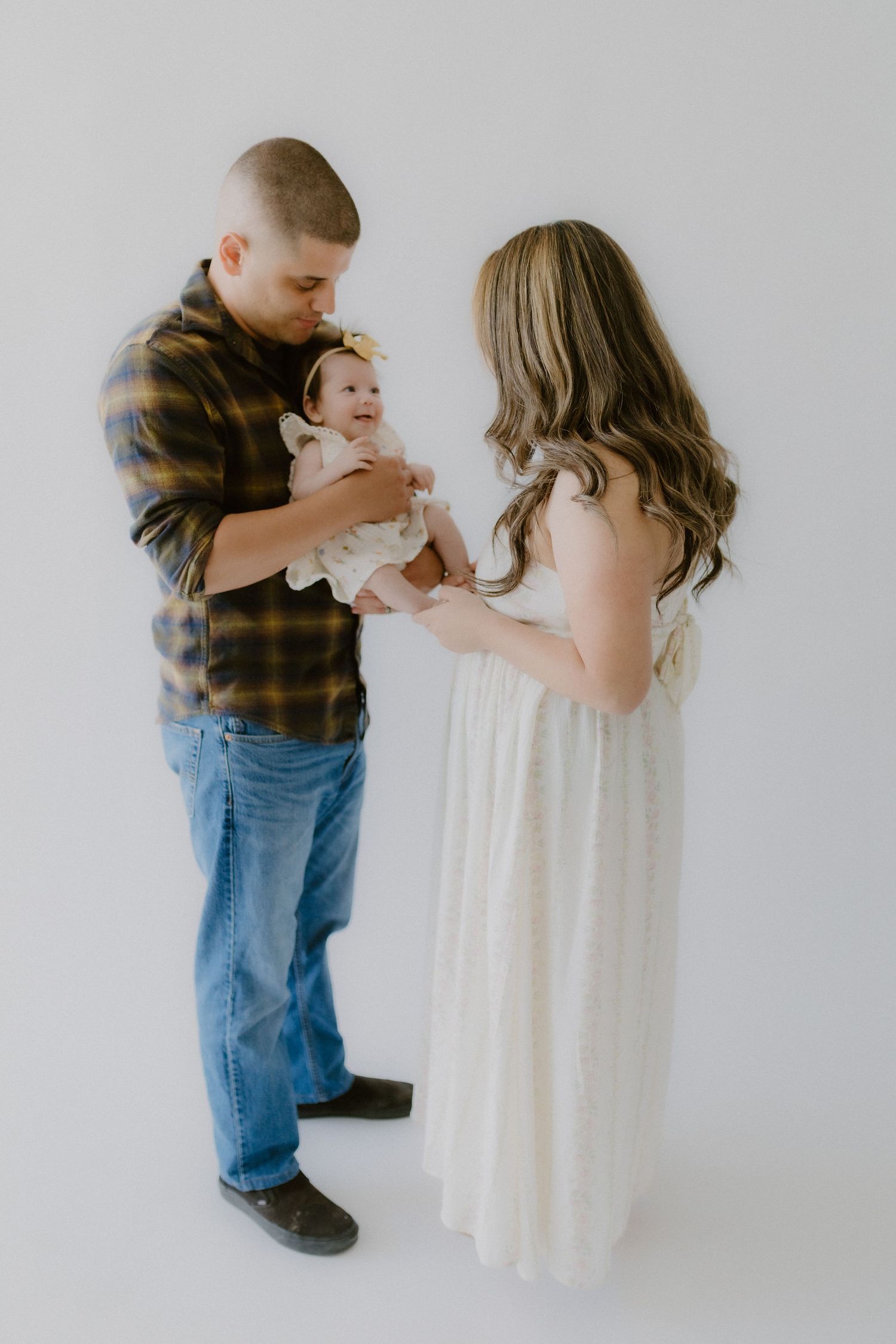 A couple in a bright studio smiles as they hold their baby girl wearing a yellow bow headband between them.