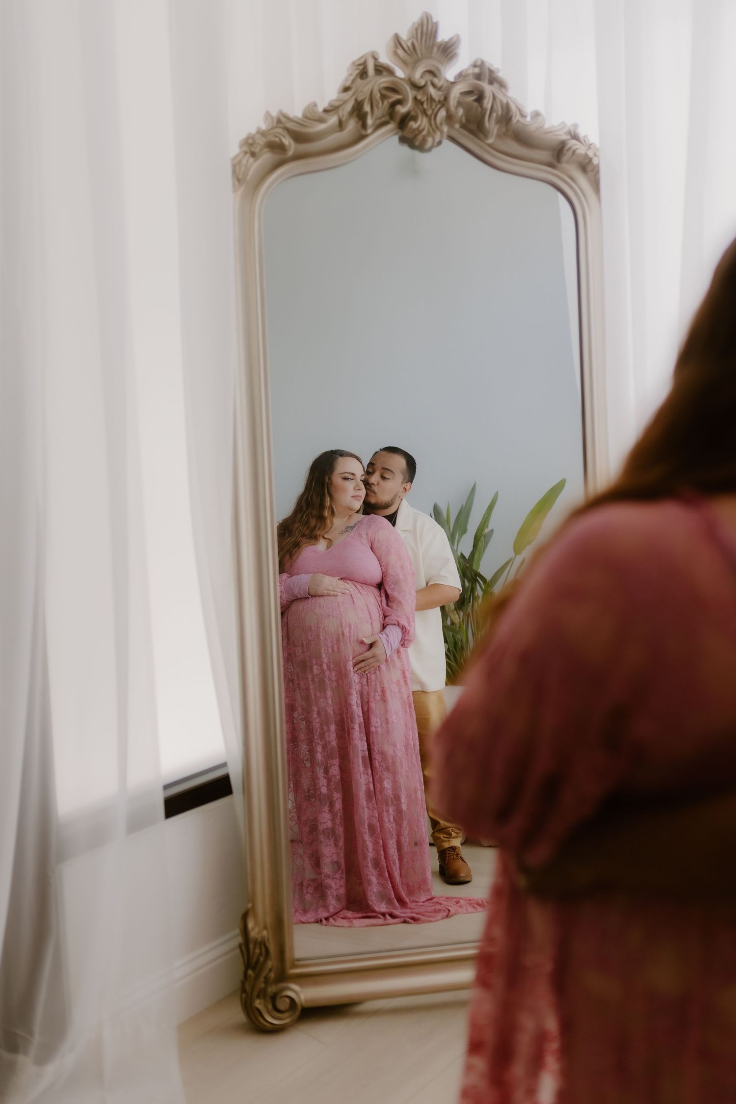 Pregnant couple reflected in ornate vintage mirror, woman in pink lace gown embraced by partner.