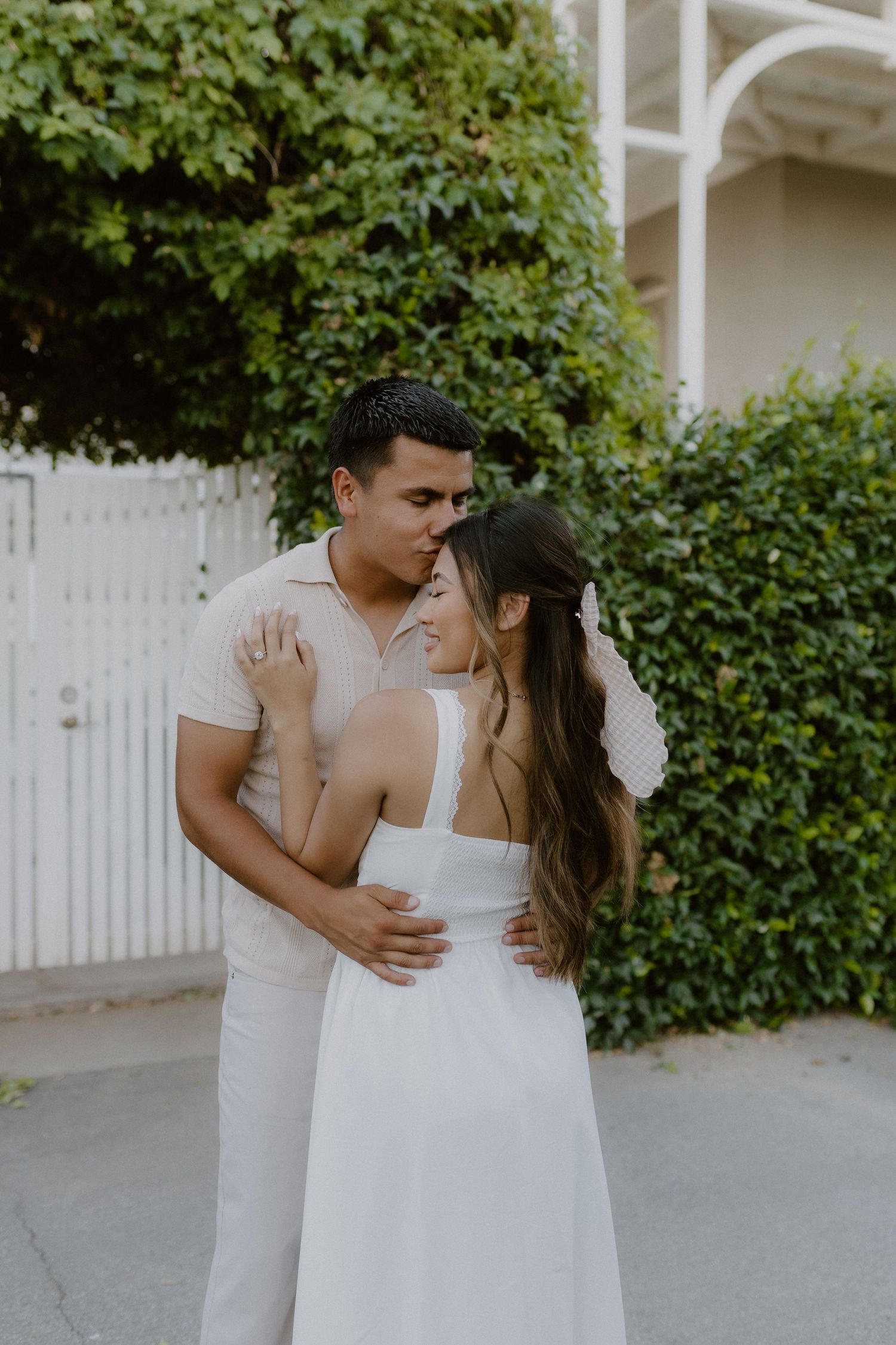 Couple in white embracing tenderly in front of lush green ivy wall and white gate.