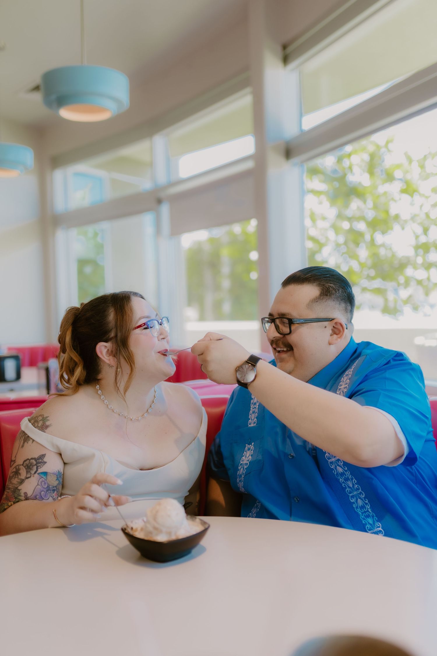 Couple sharing ice cream at retro diner booth, man in blue shirt feeding woman in off-shoulder dress.