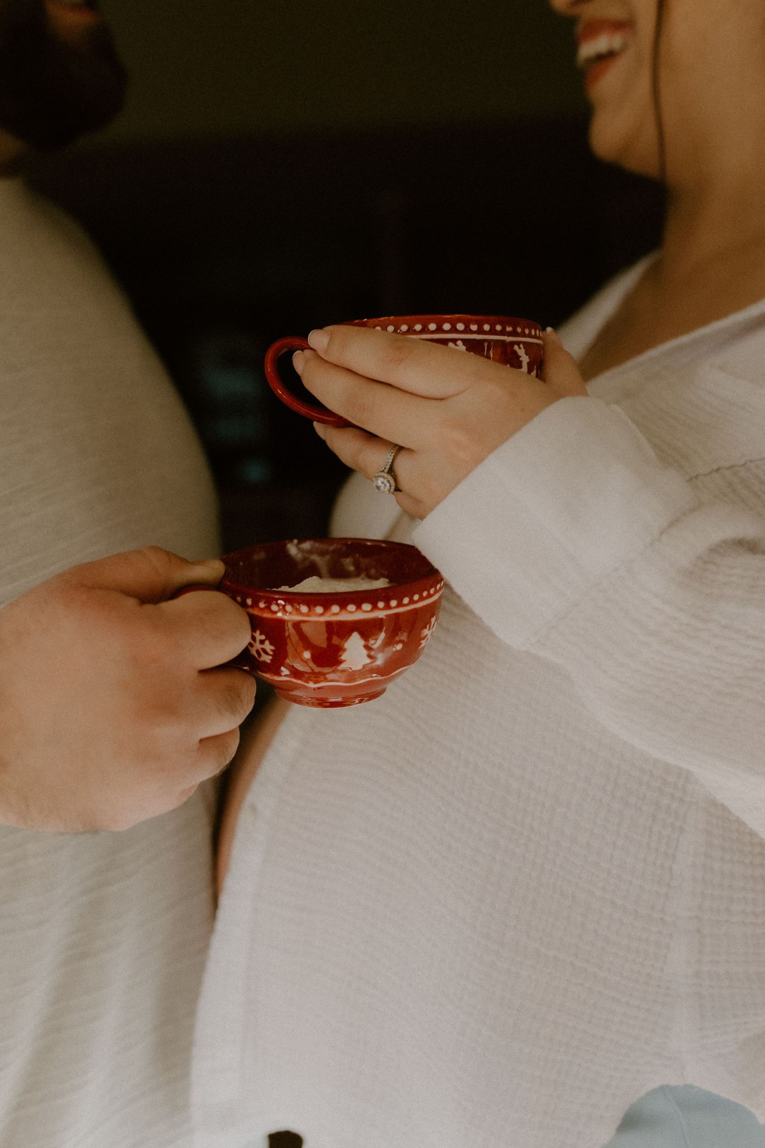 Close-up of couple holding festive red Christmas mugs together, woman showing engagement ring on hand.