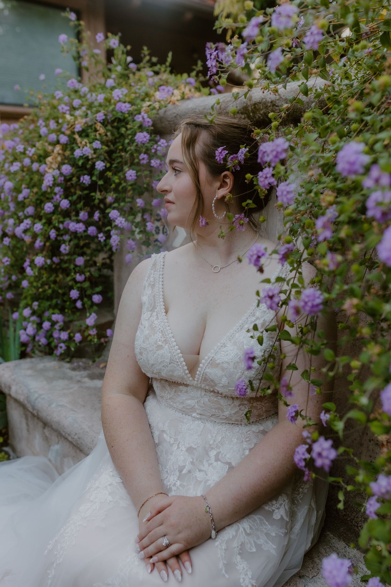 Bride in V-neck lace wedding gown posed beside cascading purple flowers.