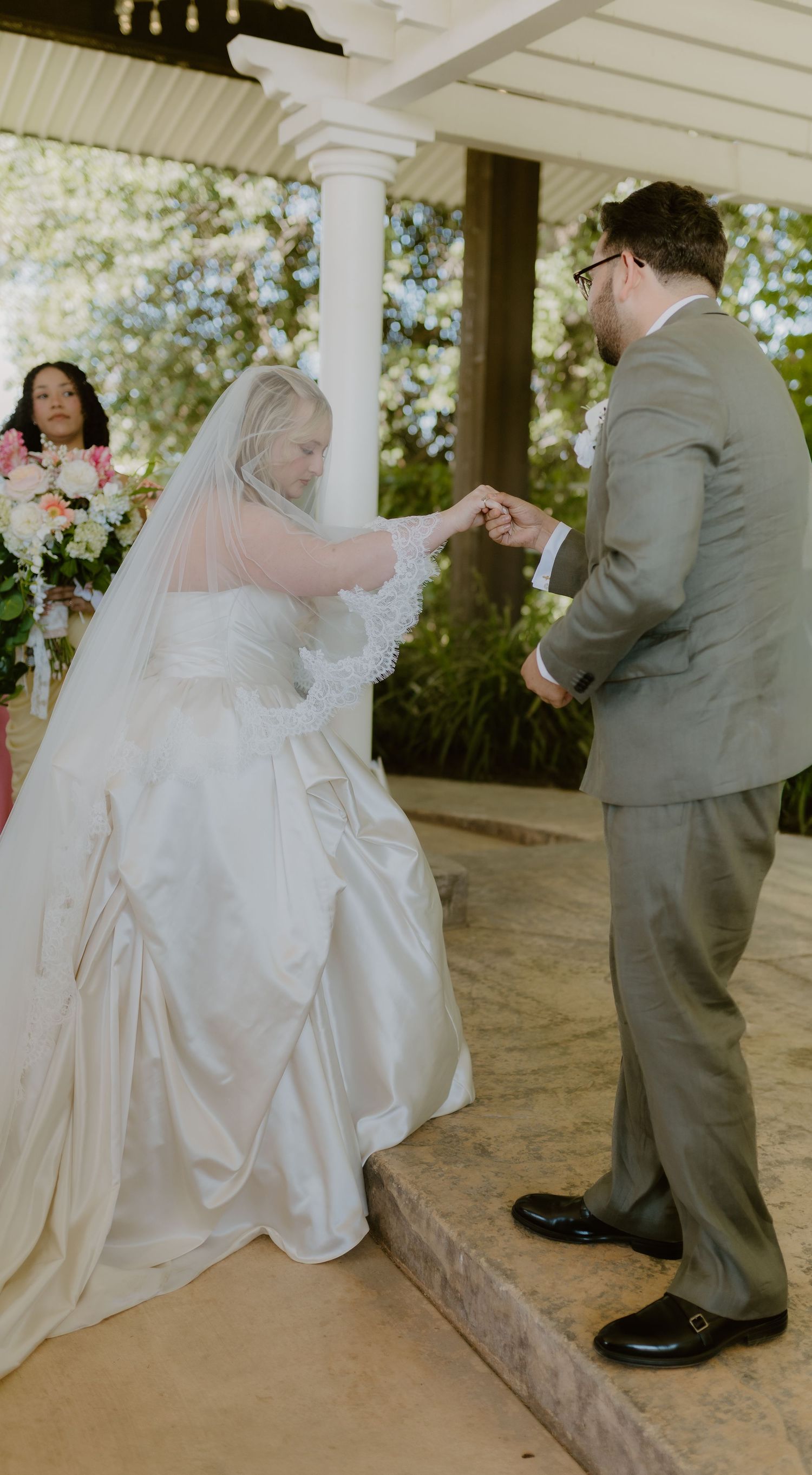 Bride in white gown and veil kneeling before groom during outdoor wedding ceremony.