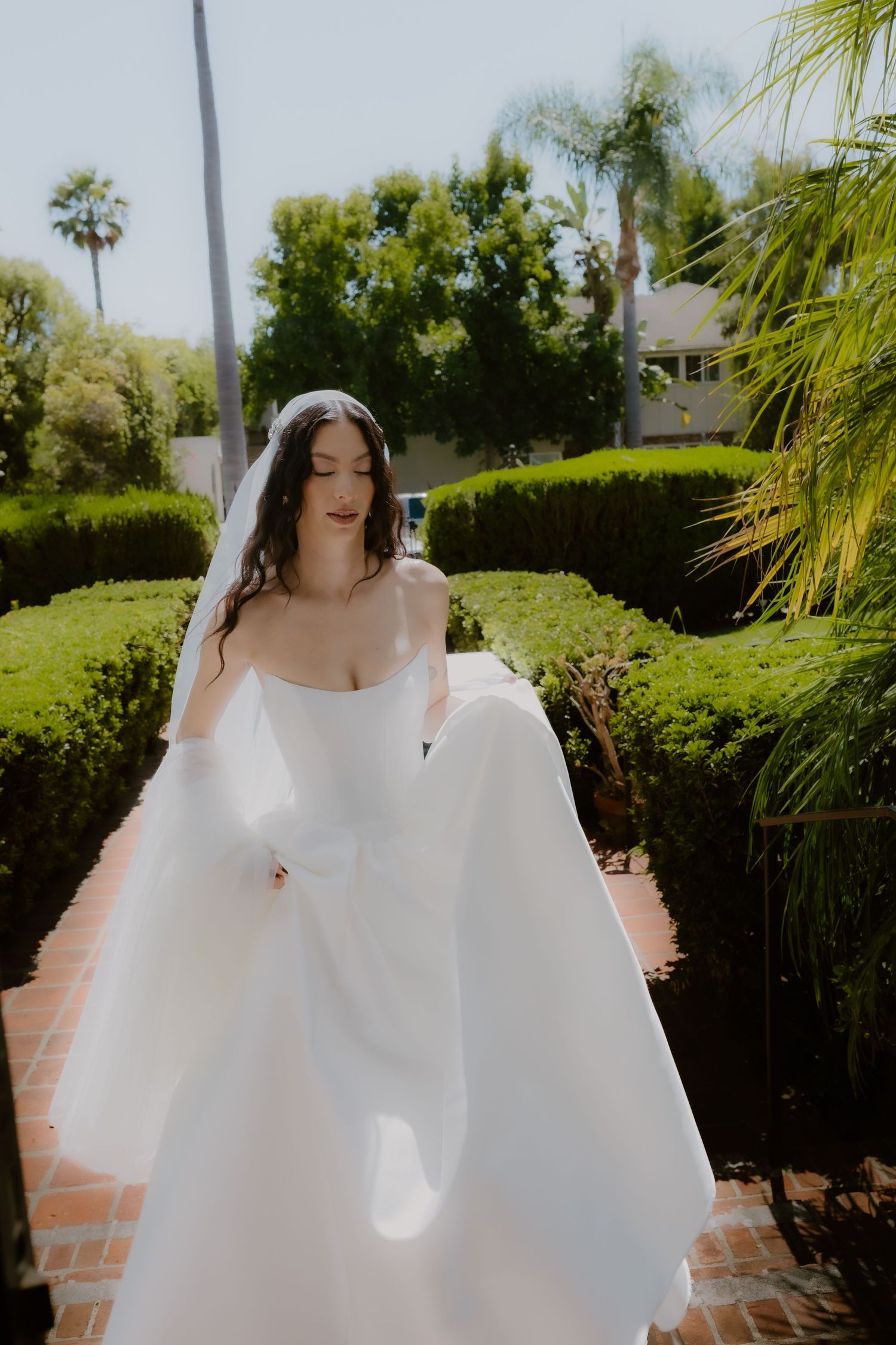 Bride in strapless white ball gown and veil walks through lush garden pathway on sunny wedding day.