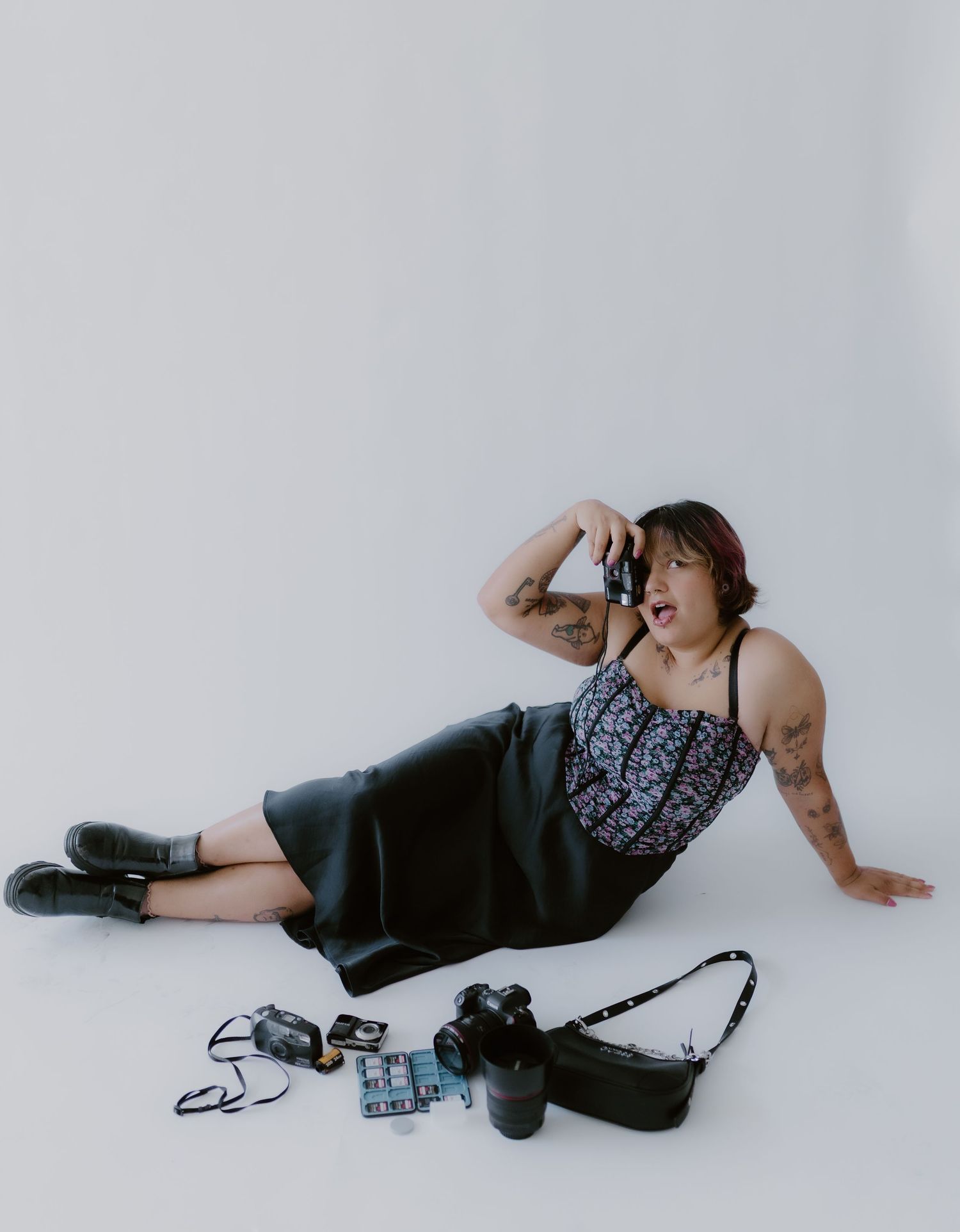 Tattooed photographer lying on studio floor holding camera, surrounded by photography equipment, wearing floral top and black skirt.