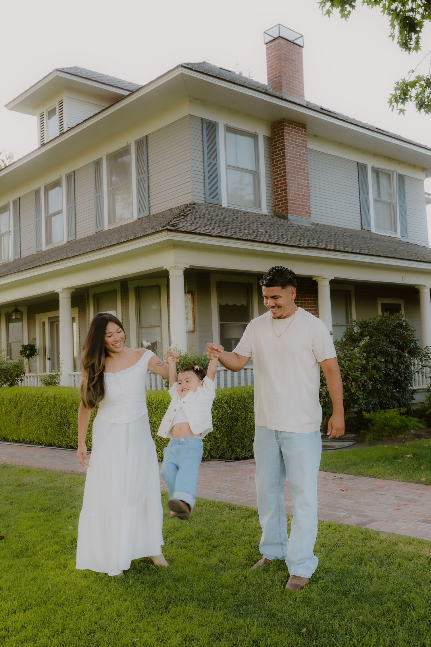 Joyful family swinging baby boy between parents on grass in front of large traditional house. The Charm House, Hanford, California