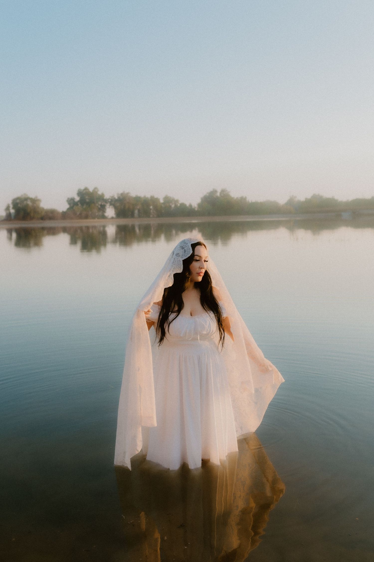 Bride in white gown and veil standing in calm lake water at golden hour.