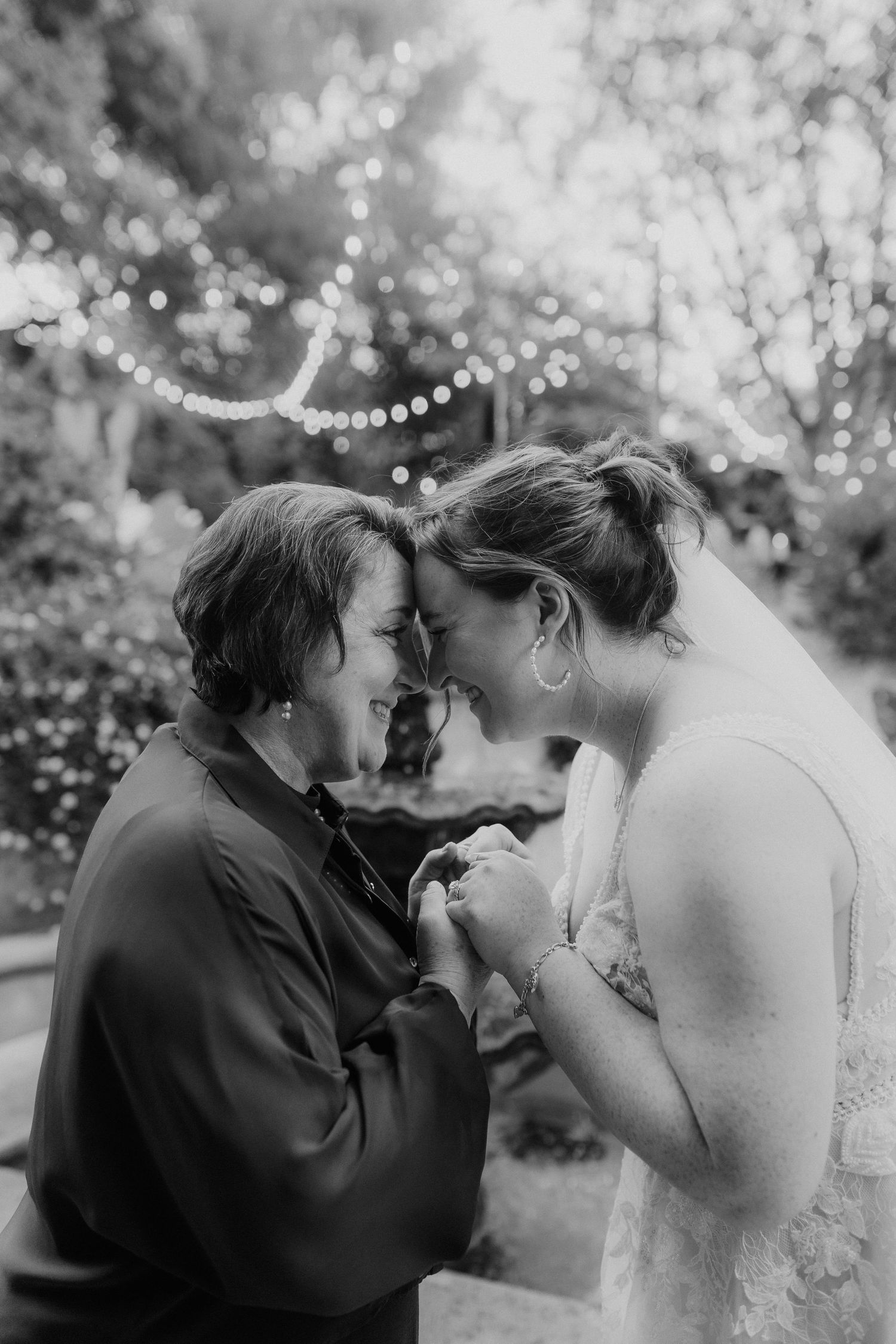 Black and white photo of two women sharing an intimate moment, touching foreheads and holding hands at an outdoor wedding.