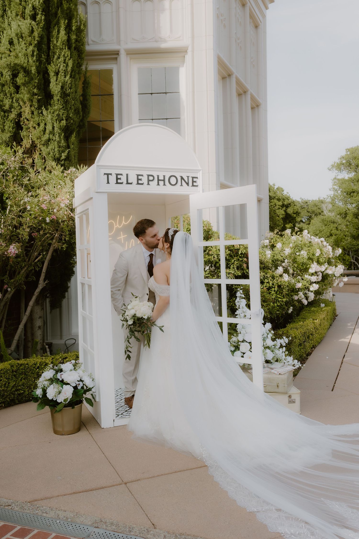 Wedding couple shares tender kiss inside white telephone booth surrounded by lush greenery and flowers.