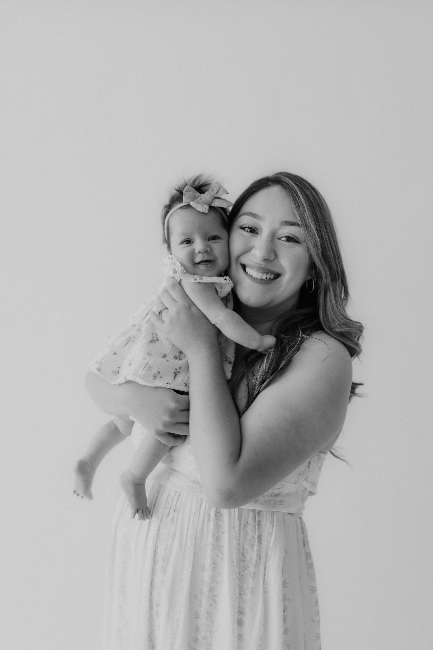 Black and white photo of a smiling young woman in a white dress holding a baby girl with a floral headband.