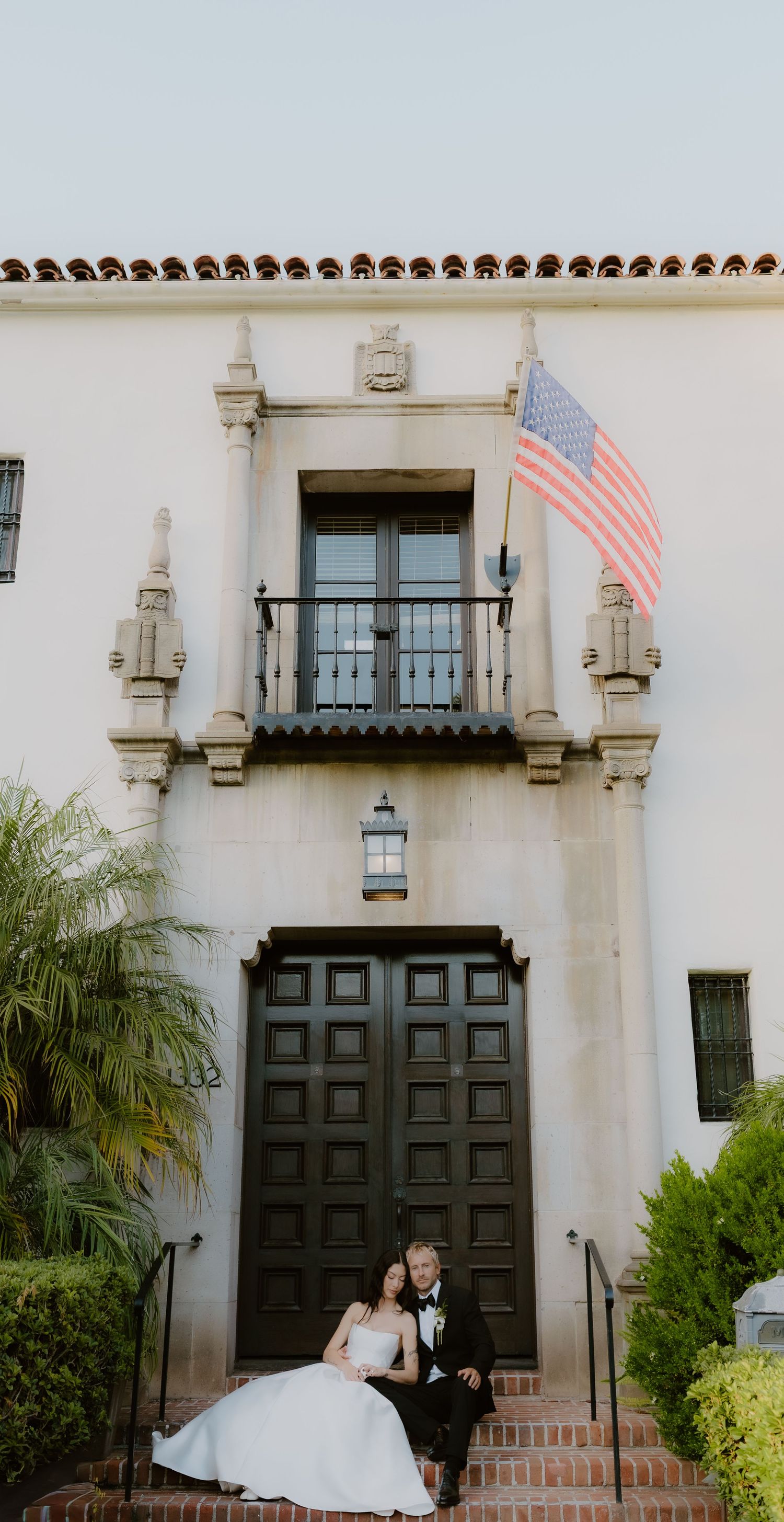 Bride and groom sit on brick steps before ornate dark doors of a white Spanish-style building with American flag.