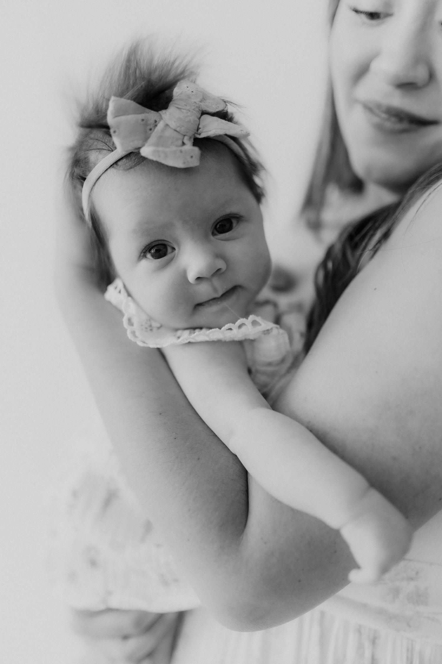 Black and white portrait of a newborn baby girl with a bow headband gazing at the camera in mothers arms.