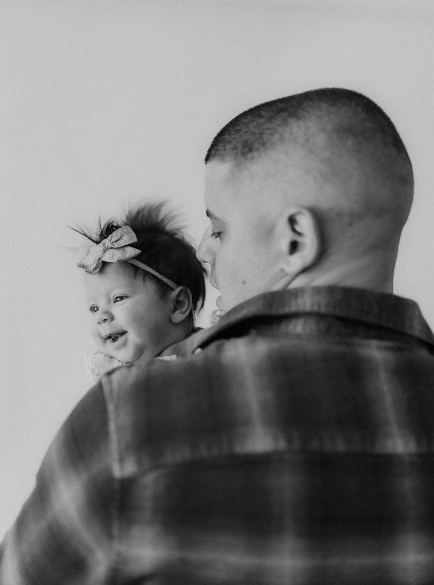 Black and white image of a father in plaid shirt holding his newborn baby girl with bow headband.