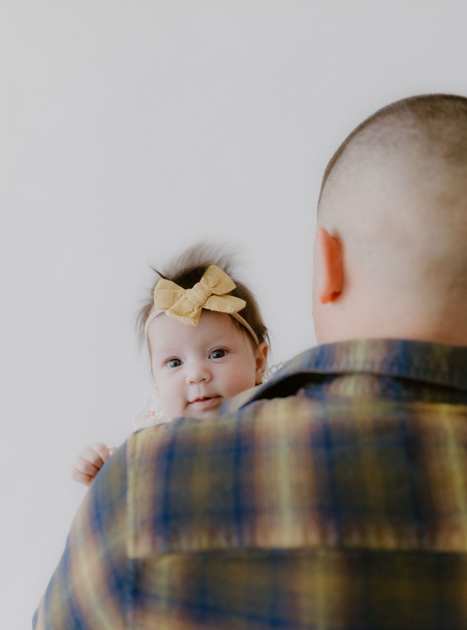 Father in plaid shirt holding baby girl with yellow bow headband who looks over his shoulder.