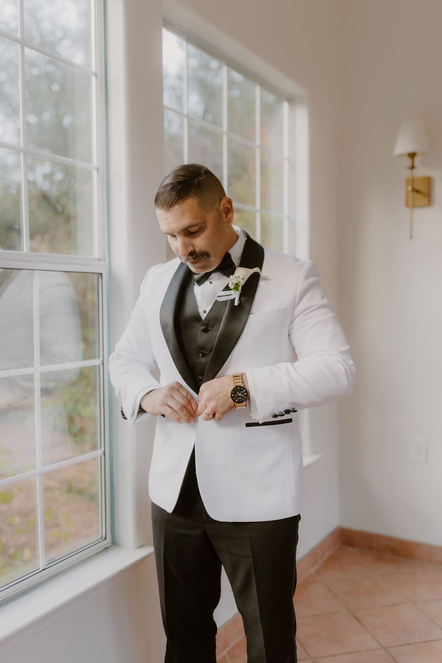 Groom in white tuxedo jacket buttons his coat while standing by large window in bright room.