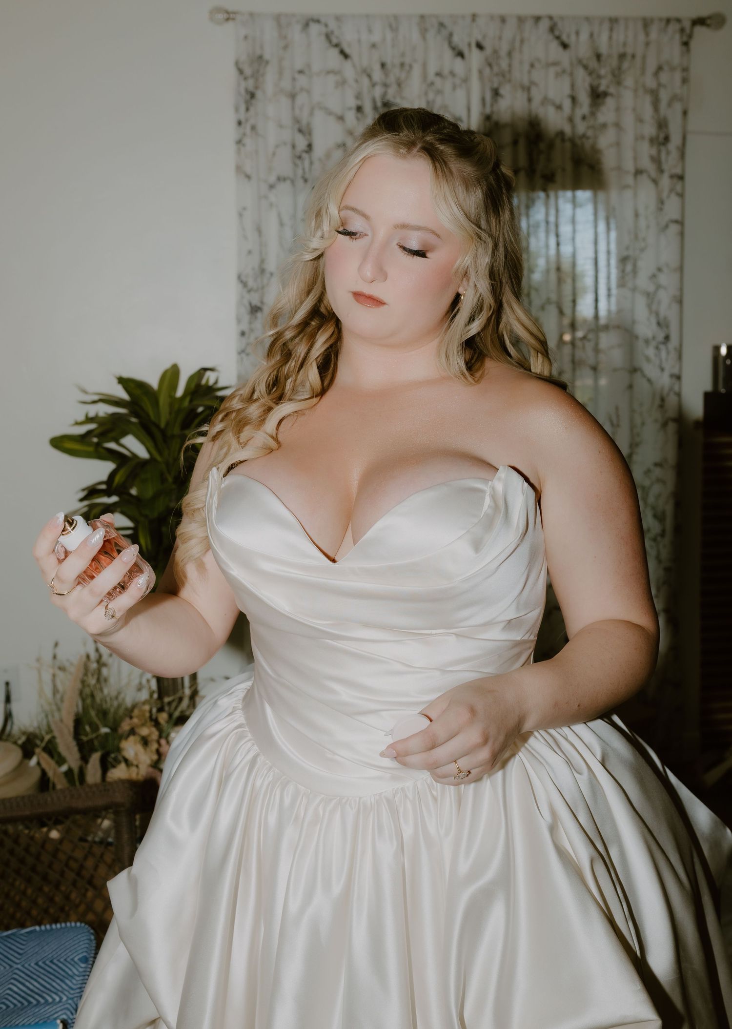 Blonde bride in strapless satin sweetheart gown holding perfume bottle while getting ready.