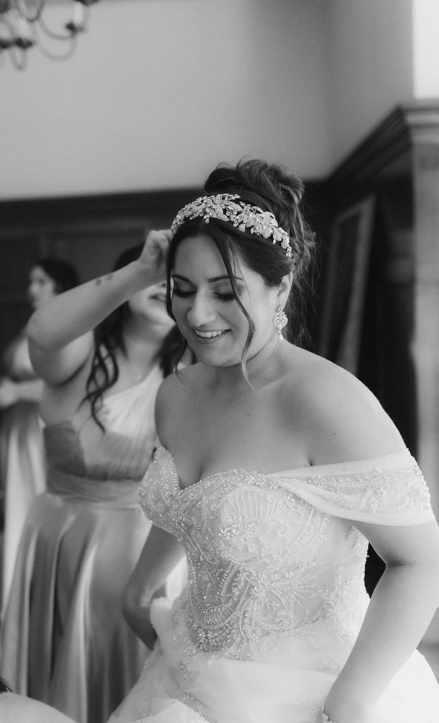Black and white photo of bride smiling as bridesmaid adjusts her crystal tiara during preparation.
