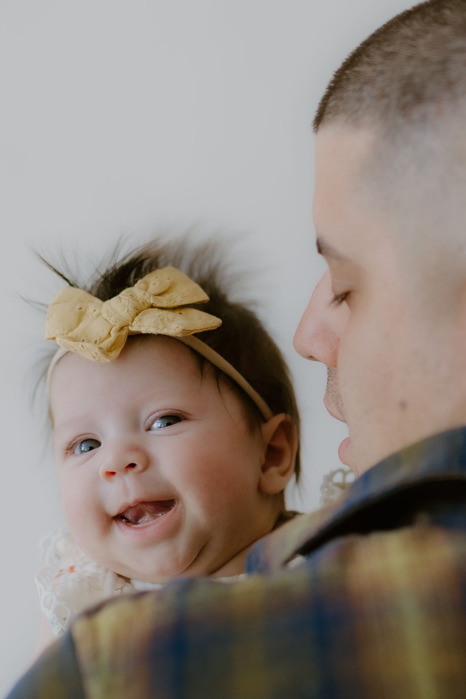 A smiling newborn baby girl with yellow bow headband being held by her father in plaid shirt.