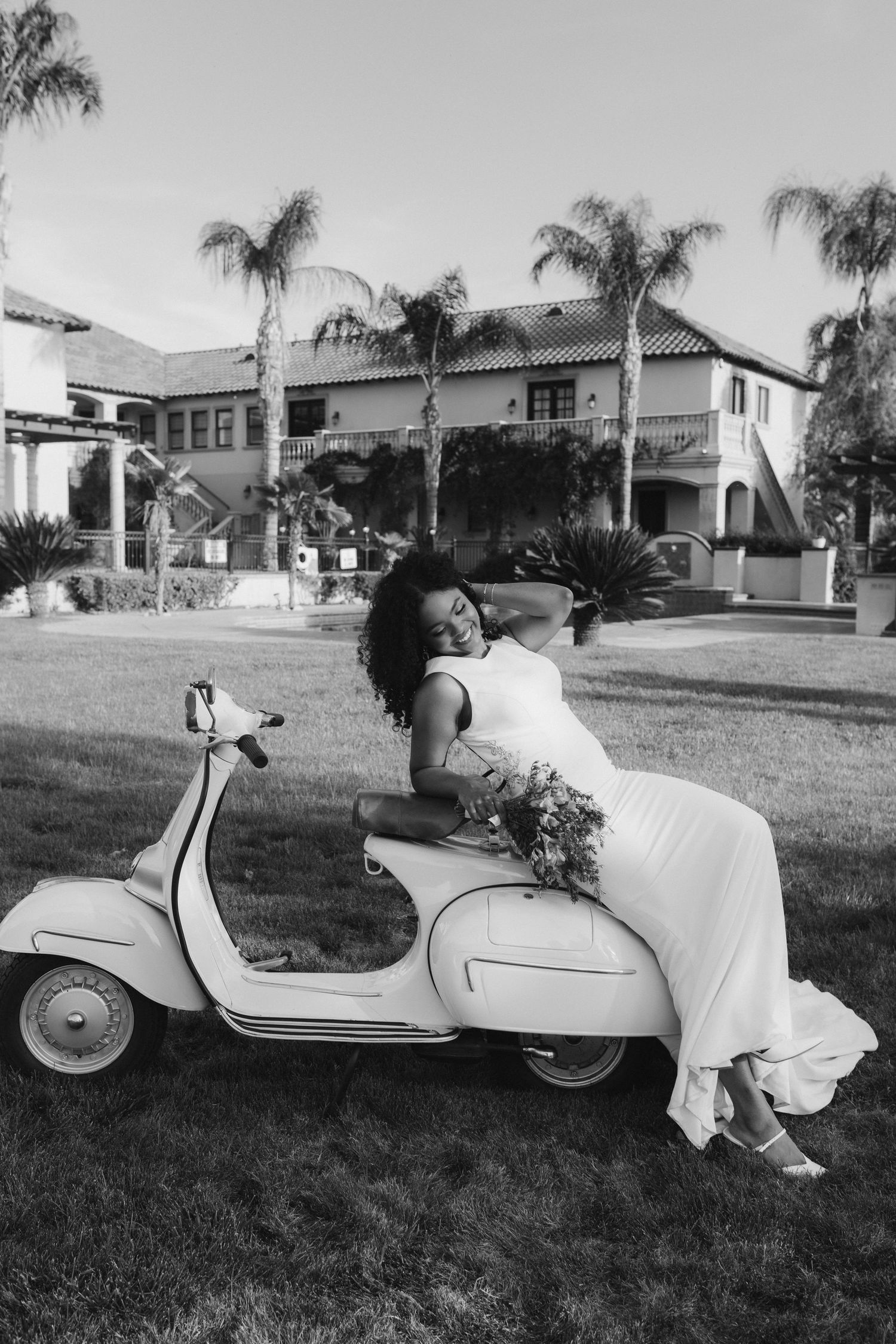 Black and white photo of a bride in a white gown leaning playfully on a vintage Vespa scooter outdoors.