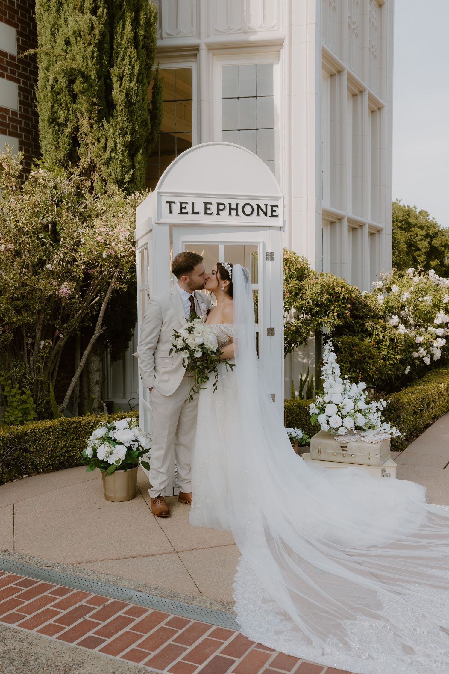 Bride and groom kiss beside white vintage telephone booth decorated with white floral arrangements.