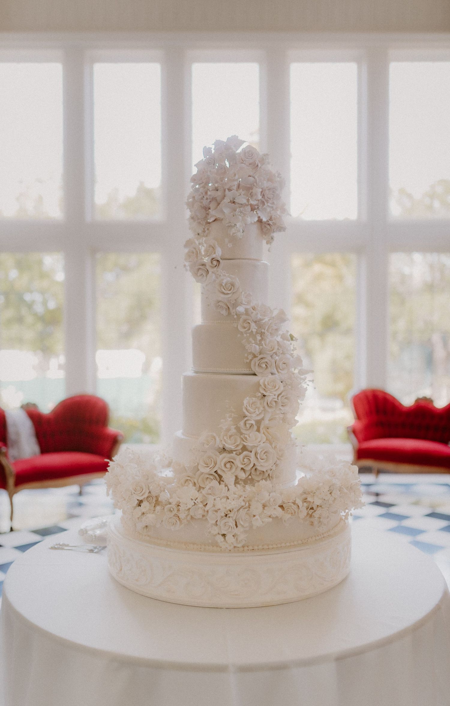 Stunning five-tier white wedding cake adorned with cascading sugar roses displayed in bright sunlit room.
