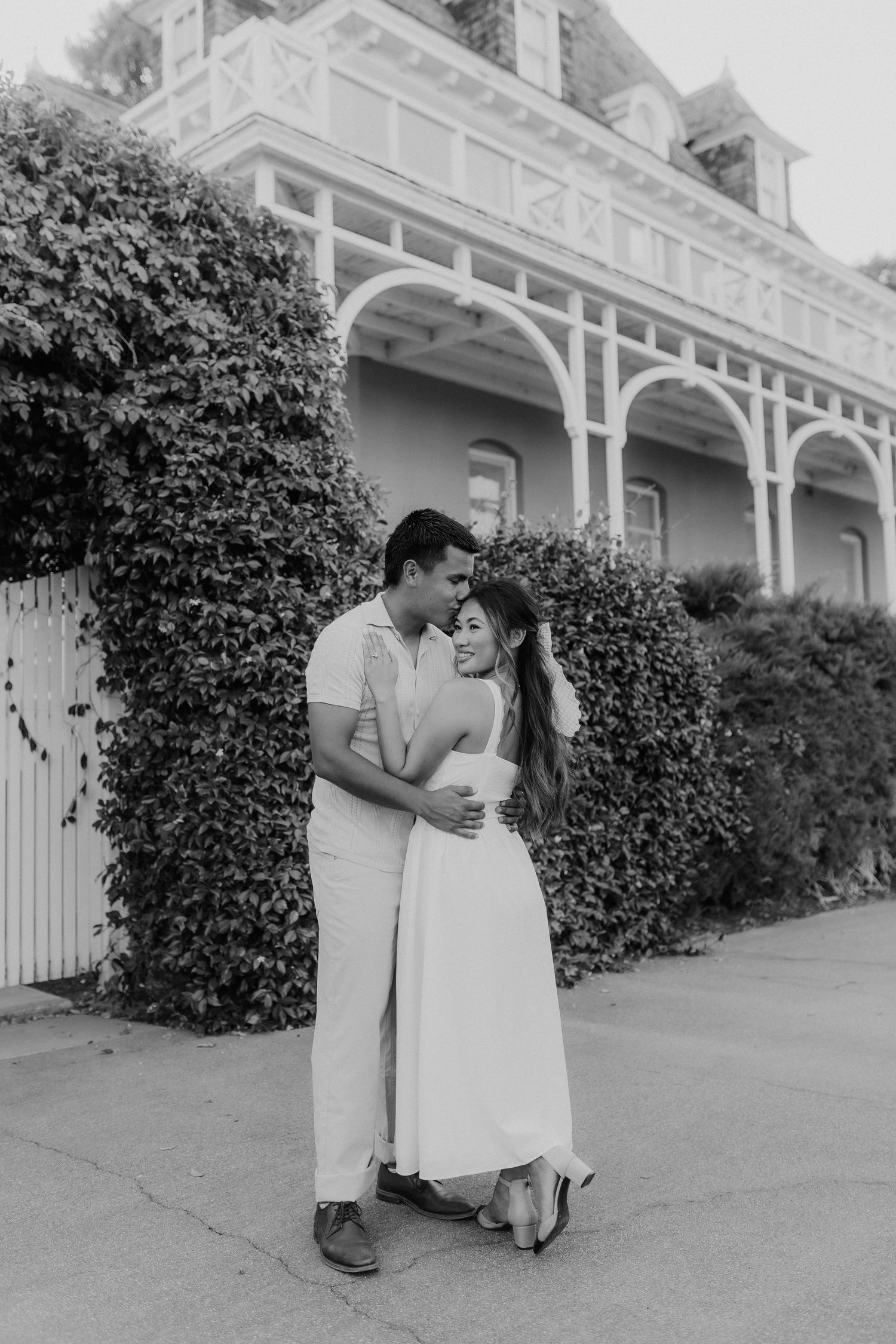 Black and white photo of couple embracing in front of ivy-covered Victorian mansion.
