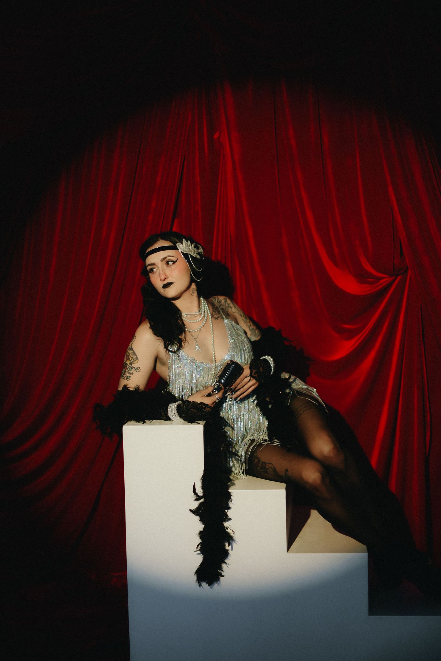 Tattooed woman in vintage flapper dress poses on white pedestal before red curtains.