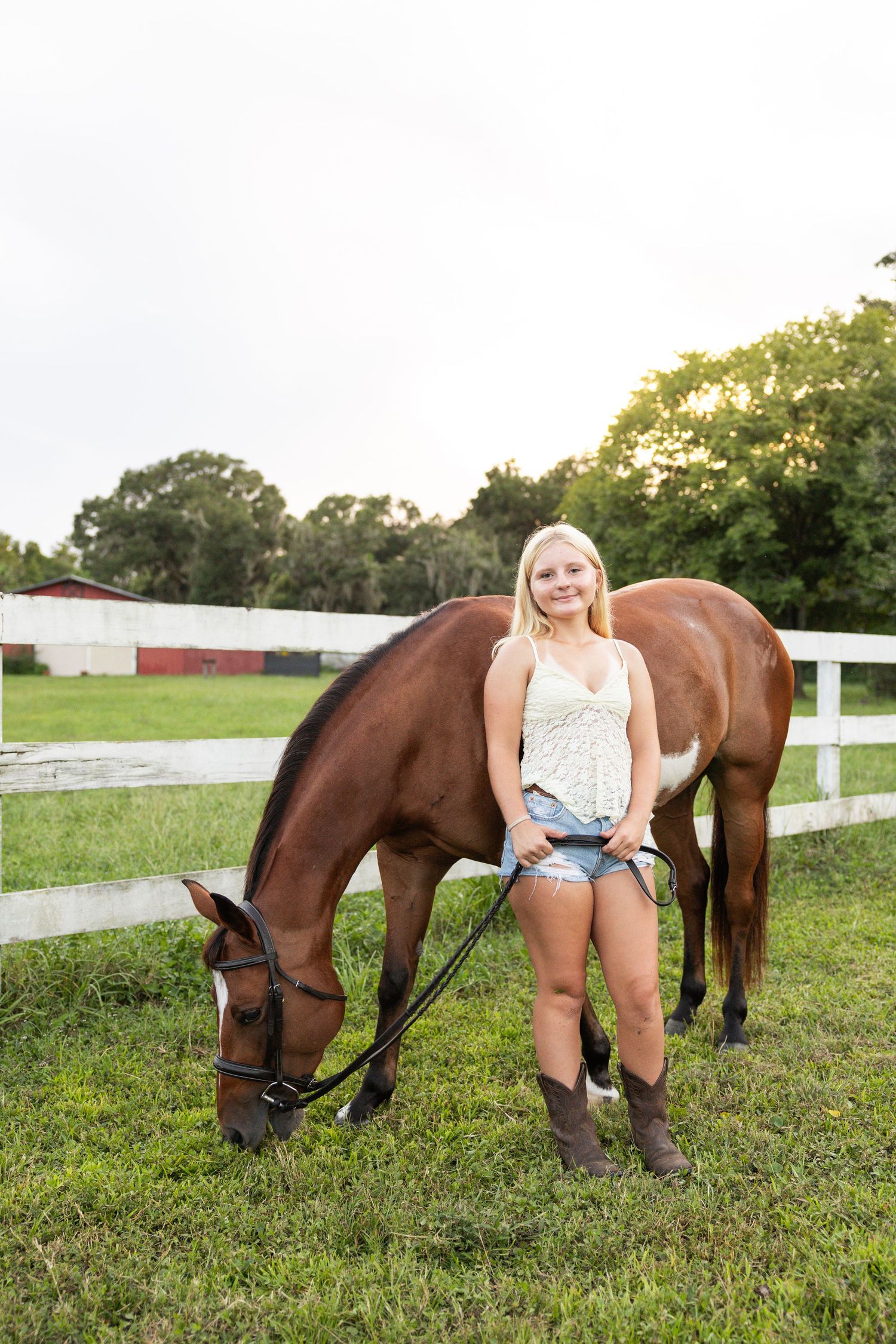 Young blonde woman in denim shorts and cowboy boots stands beside a brown horse grazing in a fenced green pasture.