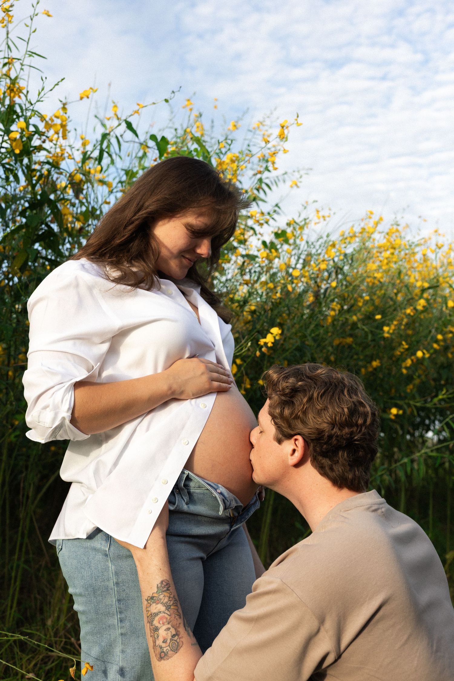 A man kisses a pregnant woman's belly outdoors among yellow wildflowers under a blue sky.