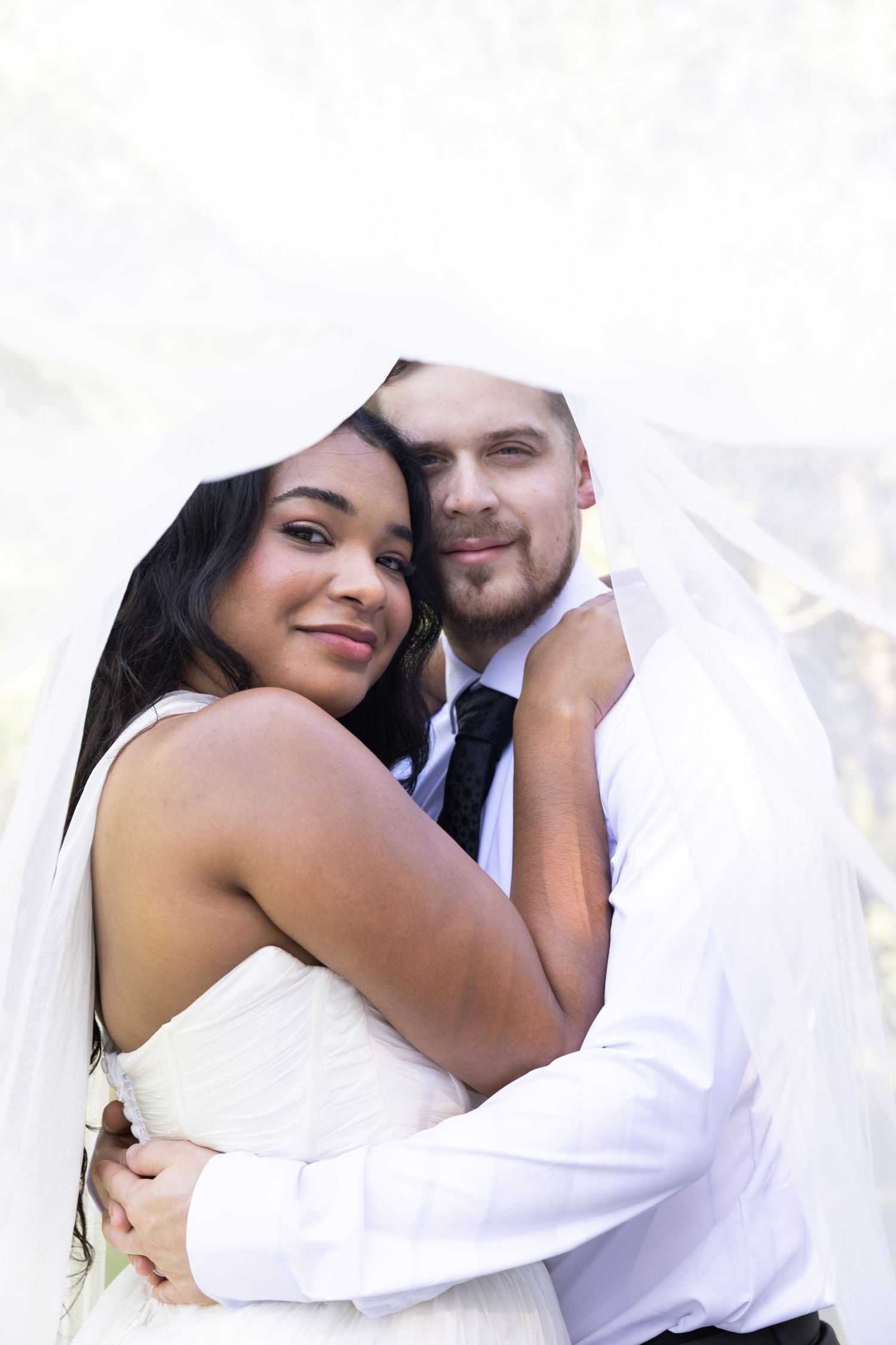 Interracial newlywed couple embracing under bridal veil, bride in white dress hugging groom in white shirt and black tie.