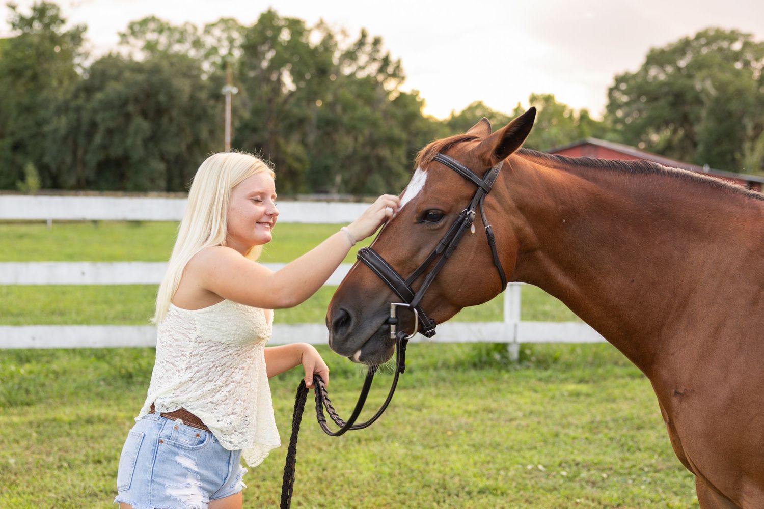 Smiling blonde woman petting a chestnut horse with a bridle in a green pasture with white fence.