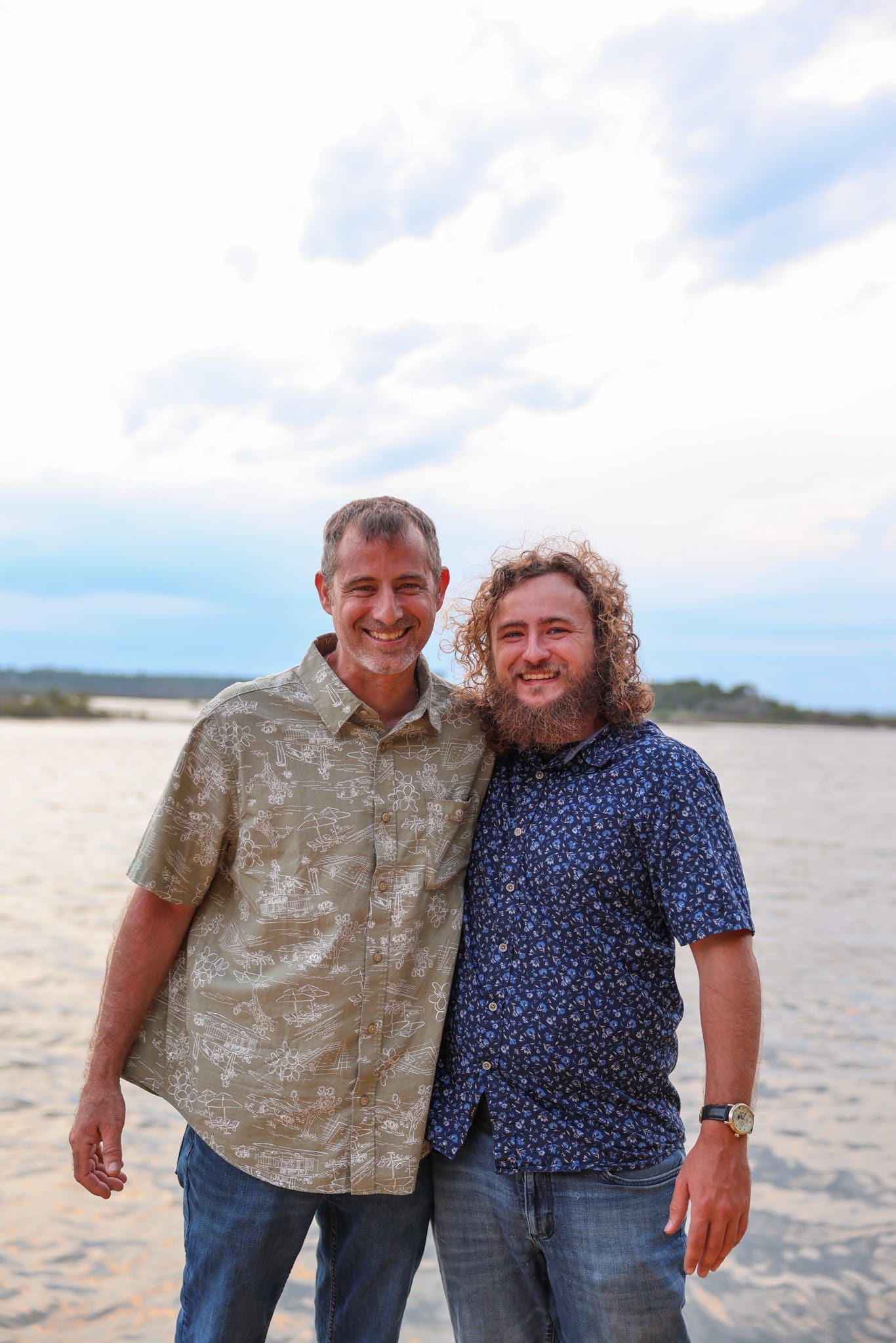 Two smiling men pose together on a sandy beach with ocean and cloudy sky in the background.