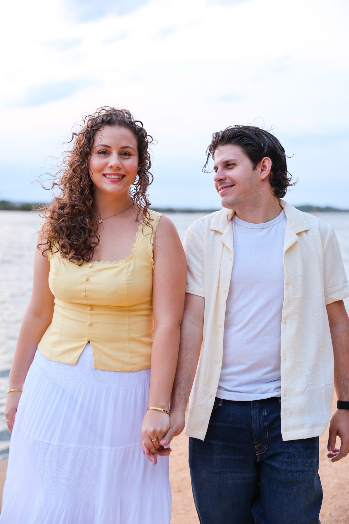 A smiling couple holds hands while walking near a waterfront, woman in yellow top and white skirt, man in jeans.
