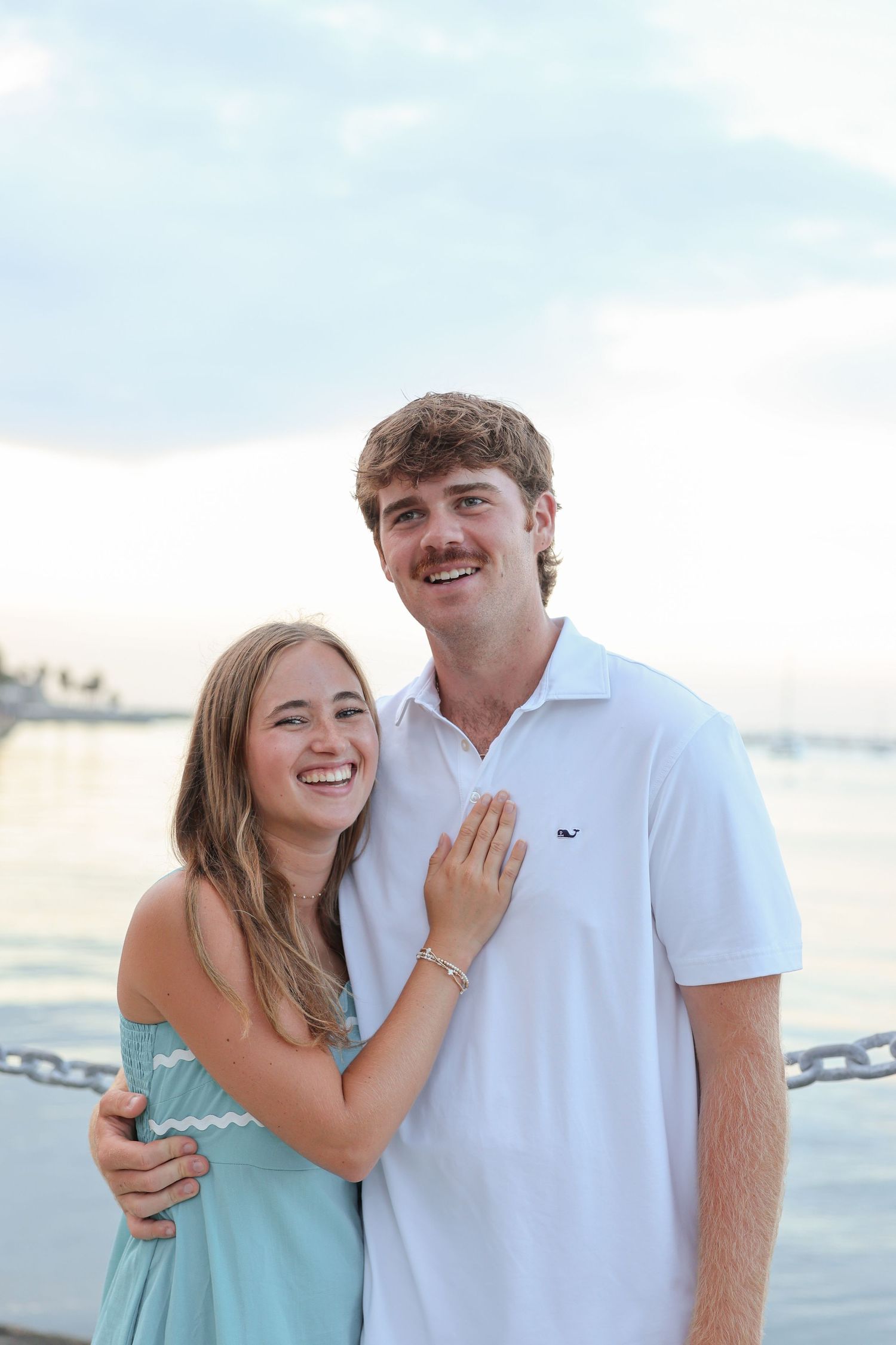 A smiling young couple embraces near the waterfront, woman in teal dress, man in white polo shirt.