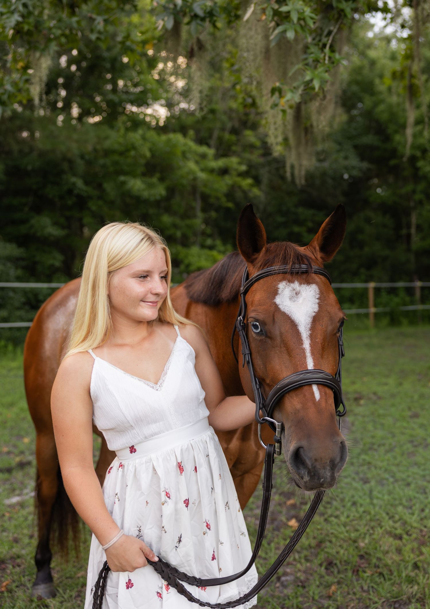 Blonde girl in white floral dress smiling beside a chestnut horse with a white blaze in a green pasture.
