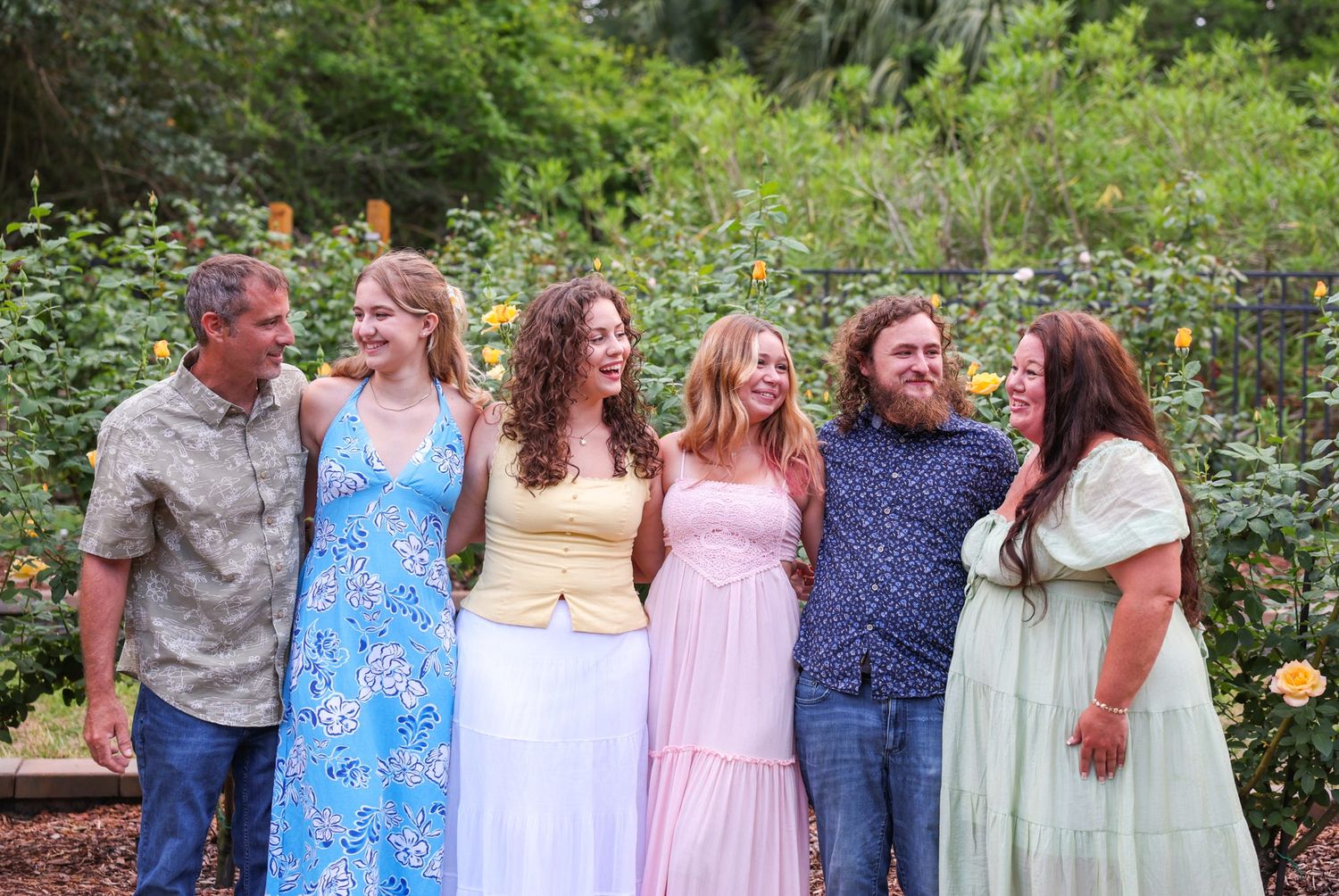 Six people smiling and laughing together in a lush garden with yellow roses, wearing pastel summer outfits.