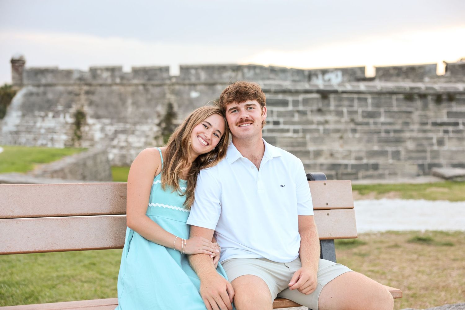 A smiling couple is sitting together on a bench in front of the historic Castillo de San Marcos fort in St. Augustine.
