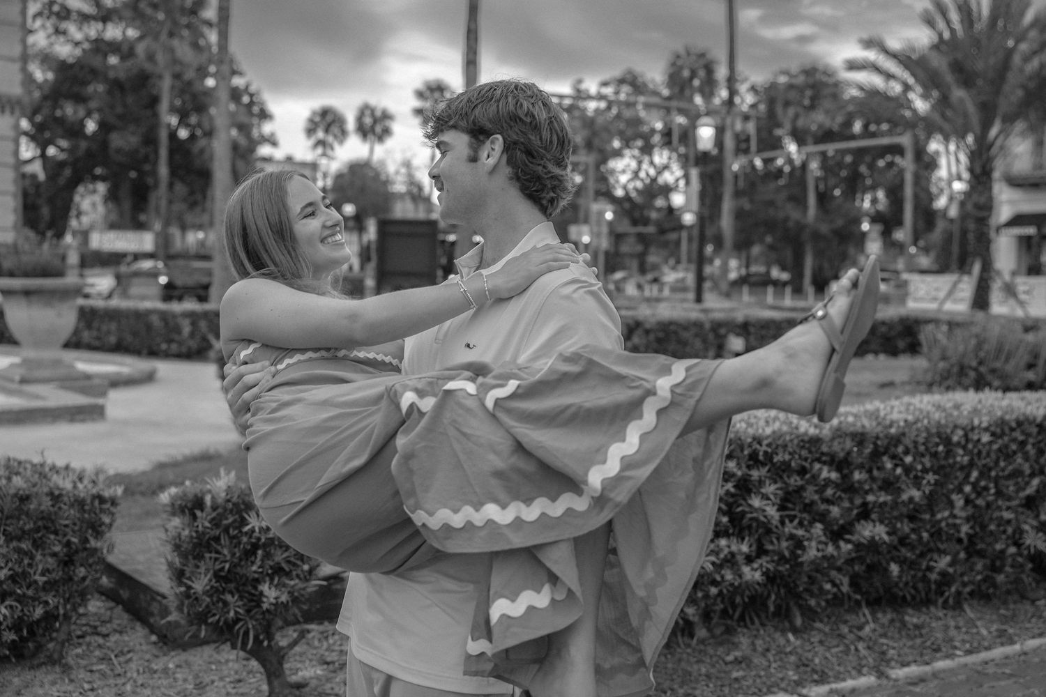 Black and white photo of a man carrying a woman in his arms outdoors near hedges and palm trees.