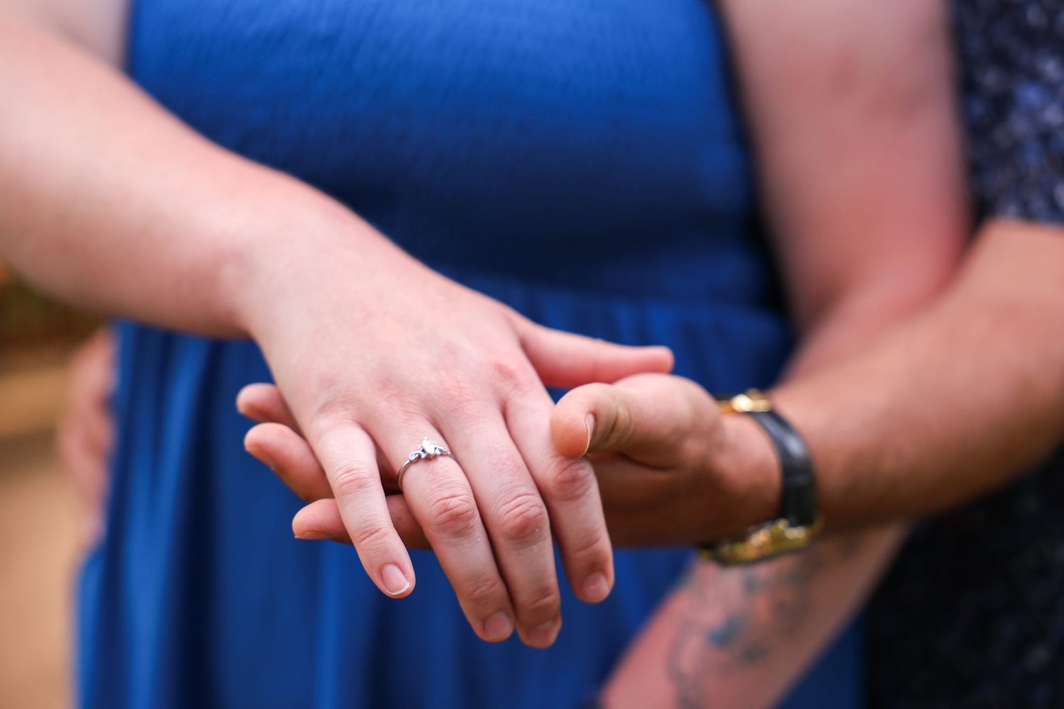 Close-up of couple holding hands showing engagement ring, woman in blue dress, man with tattoo and watch.