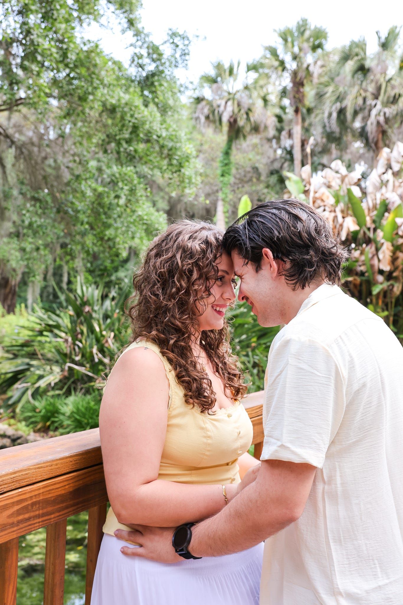 A couple shares a tender moment on a wooden deck surrounded by lush tropical greenery, the woman in yellow smiling warmly.