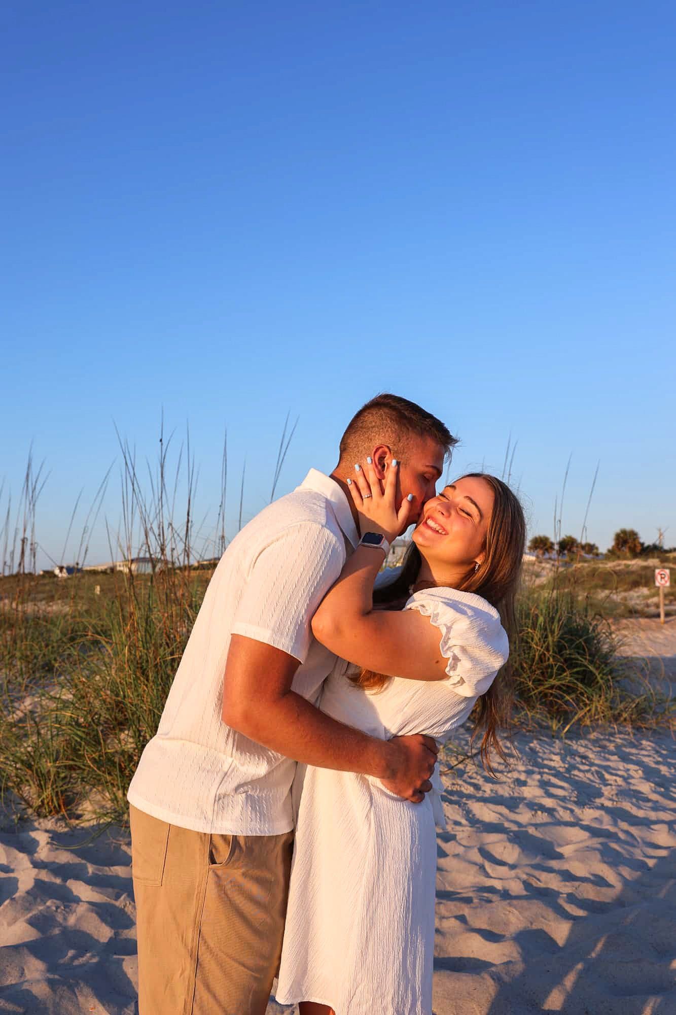 Couple embracing on sandy beach at golden hour, woman in white dress smiling, man in white shirt, blue sky background.
