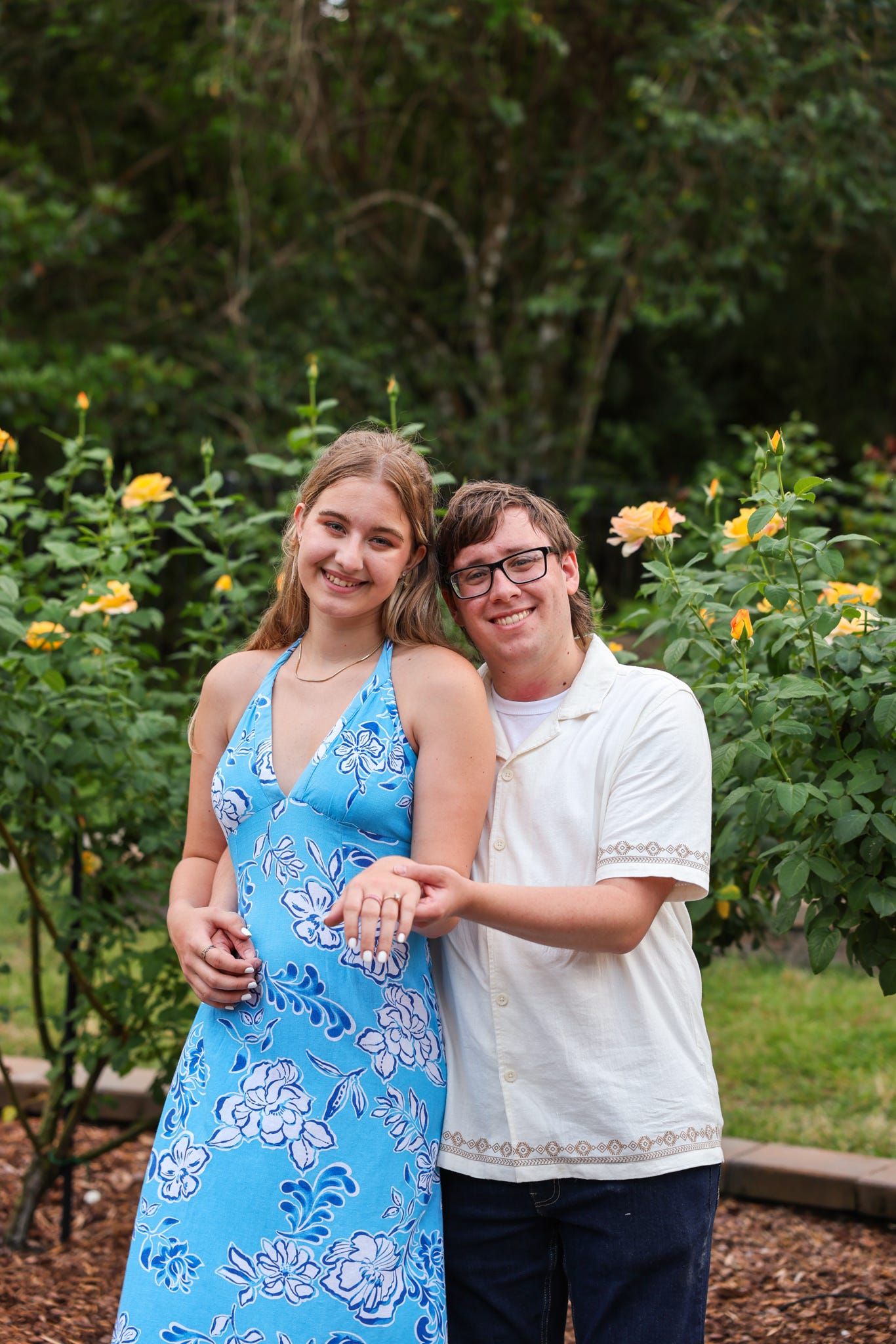 Smiling couple posing in garden with yellow roses, woman in blue floral dress, man in white shirt, showing engagement ring.