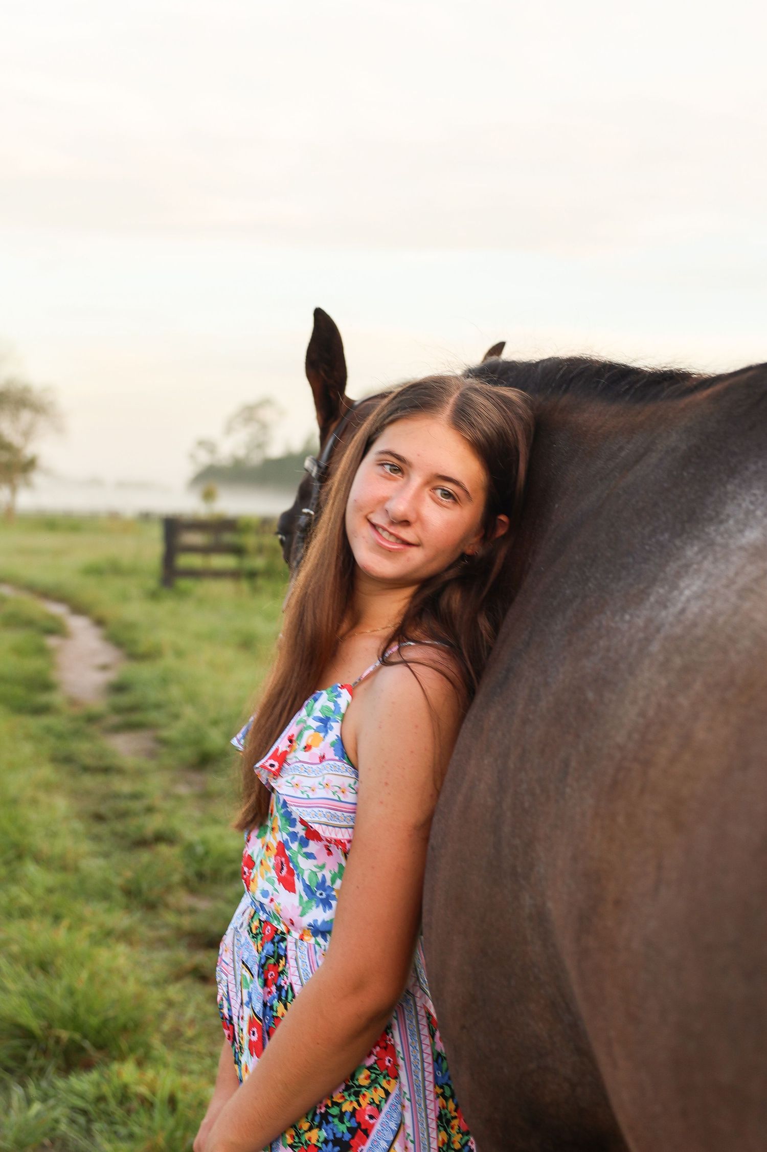 A smiling teenage girl in a floral dress leans against a dark brown horse in a green rural field at dusk.